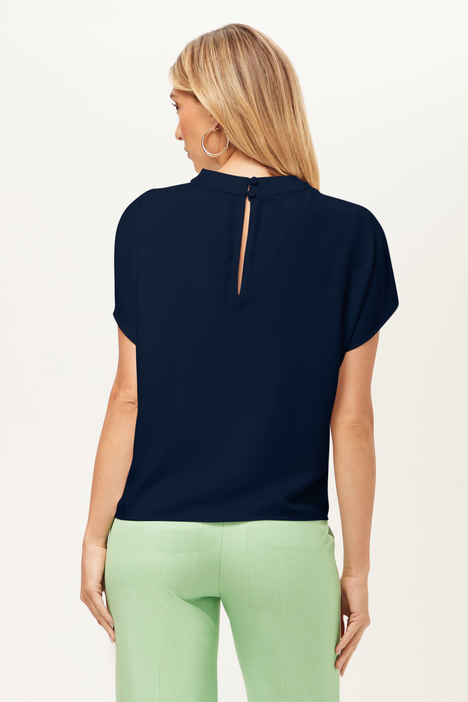 A woman with long blonde hair is shown from the back, wearing the navy Bisque Top with a keyhole button closure and light green pants. She has hoop earrings and stands against a plain, light background.