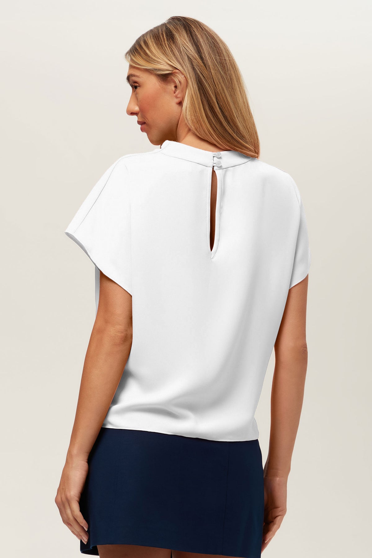 A woman with long, straight blonde hair is seen from the back wearing the Bisque Top—a white short-sleeve blouse featuring a keyhole button neckline—paired with a navy skirt against a plain light background.