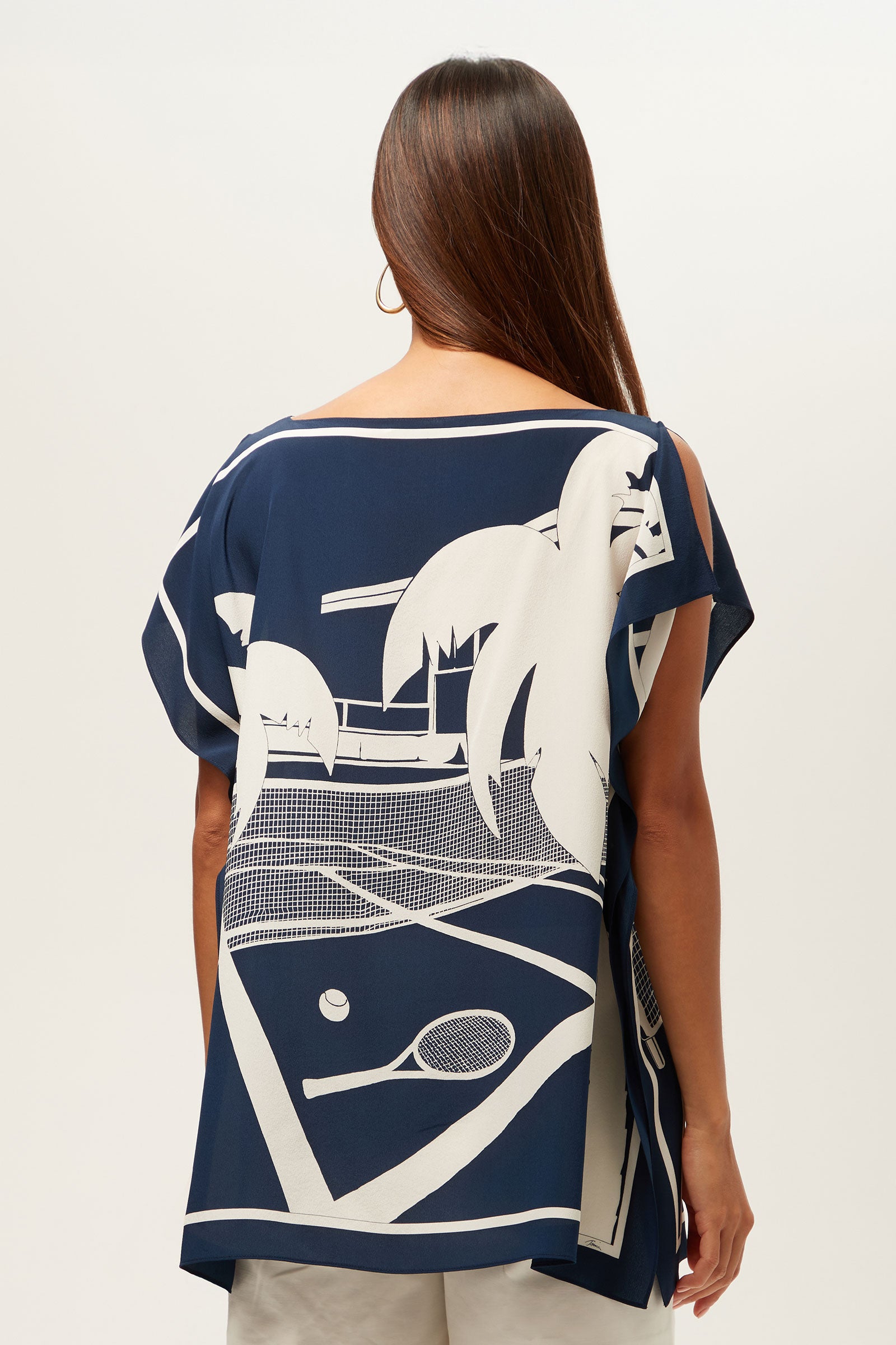 A woman with long brown hair, seen from behind, wears the Rina Split-Sleeve Top - Tennis Scarf Silk, featuring a loose navy blue fit and a white graphic of a tennis court, palm trees, net, tennis ball, and racquet. She pairs it with light shorts.