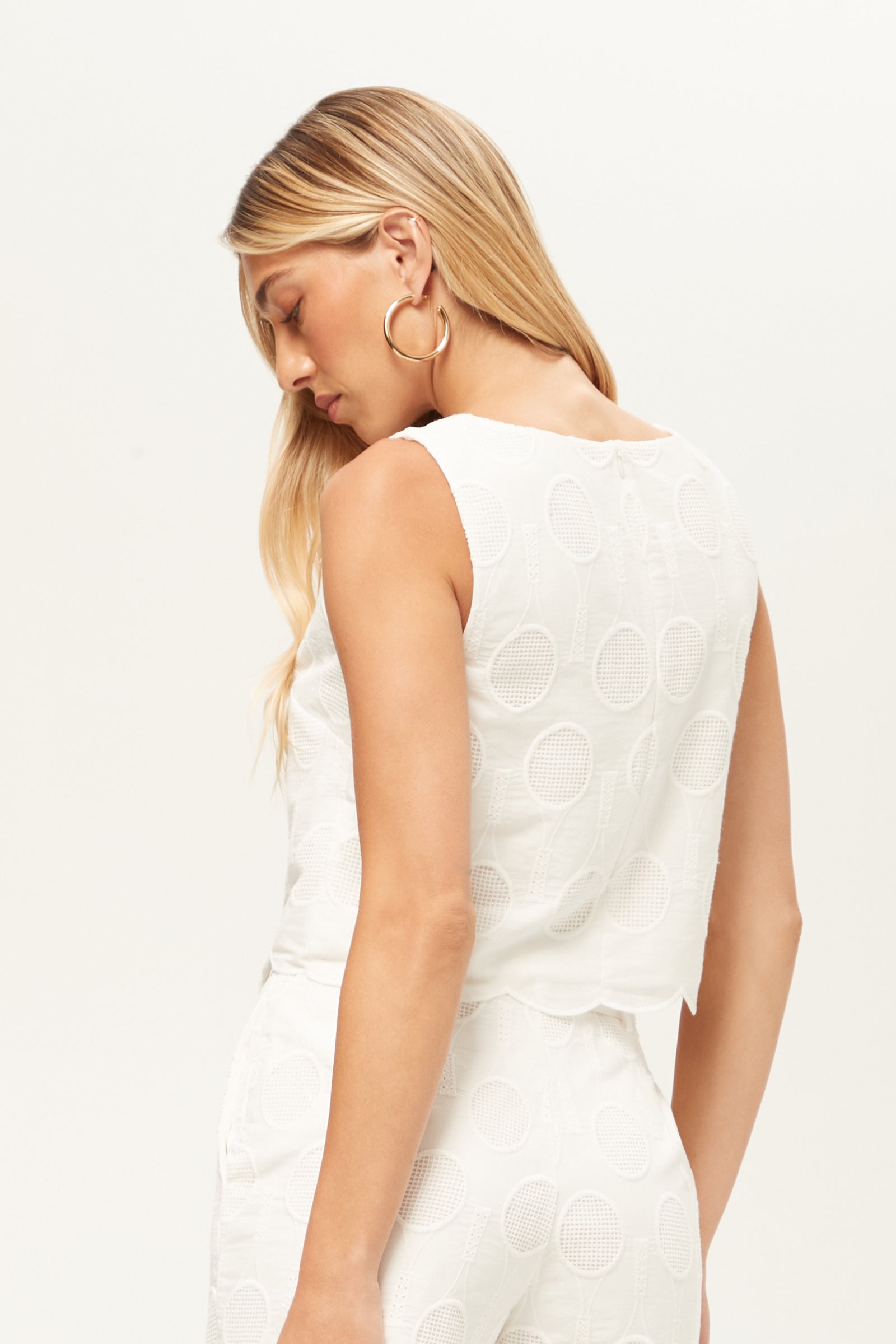 A woman with long blonde hair models the Split Sleeveless Crop Top - Tennis Eyelet, paired with matching pants featuring a subtle tennis racket pattern, as she looks down over her shoulder against a plain light background.