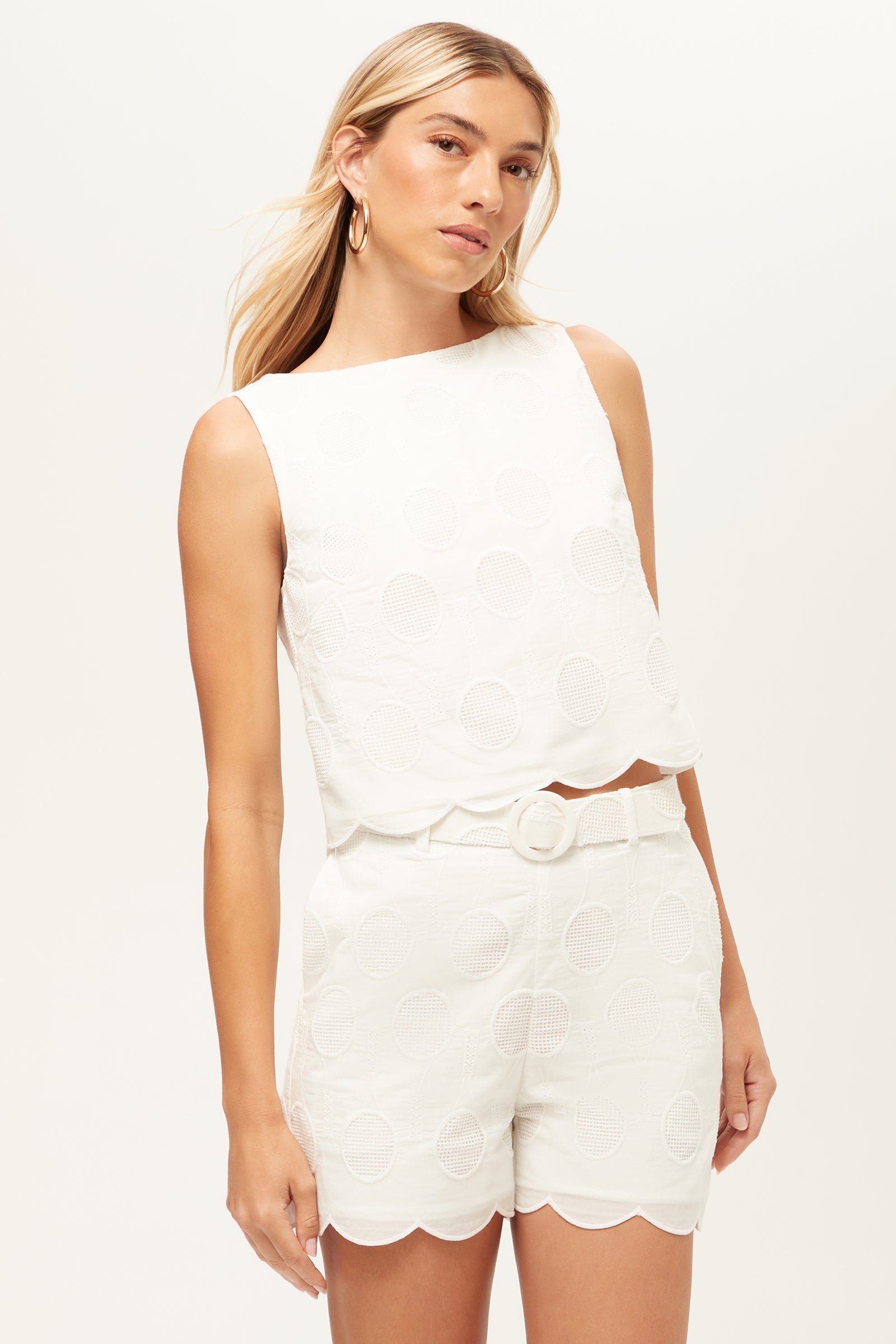 A woman with long blonde hair models the Split Sleeveless Crop Top - Tennis Eyelet in Whitewash, paired with matching high-waisted shorts featuring a subtle circular pattern and scalloped hems, posing confidently against a plain white background.