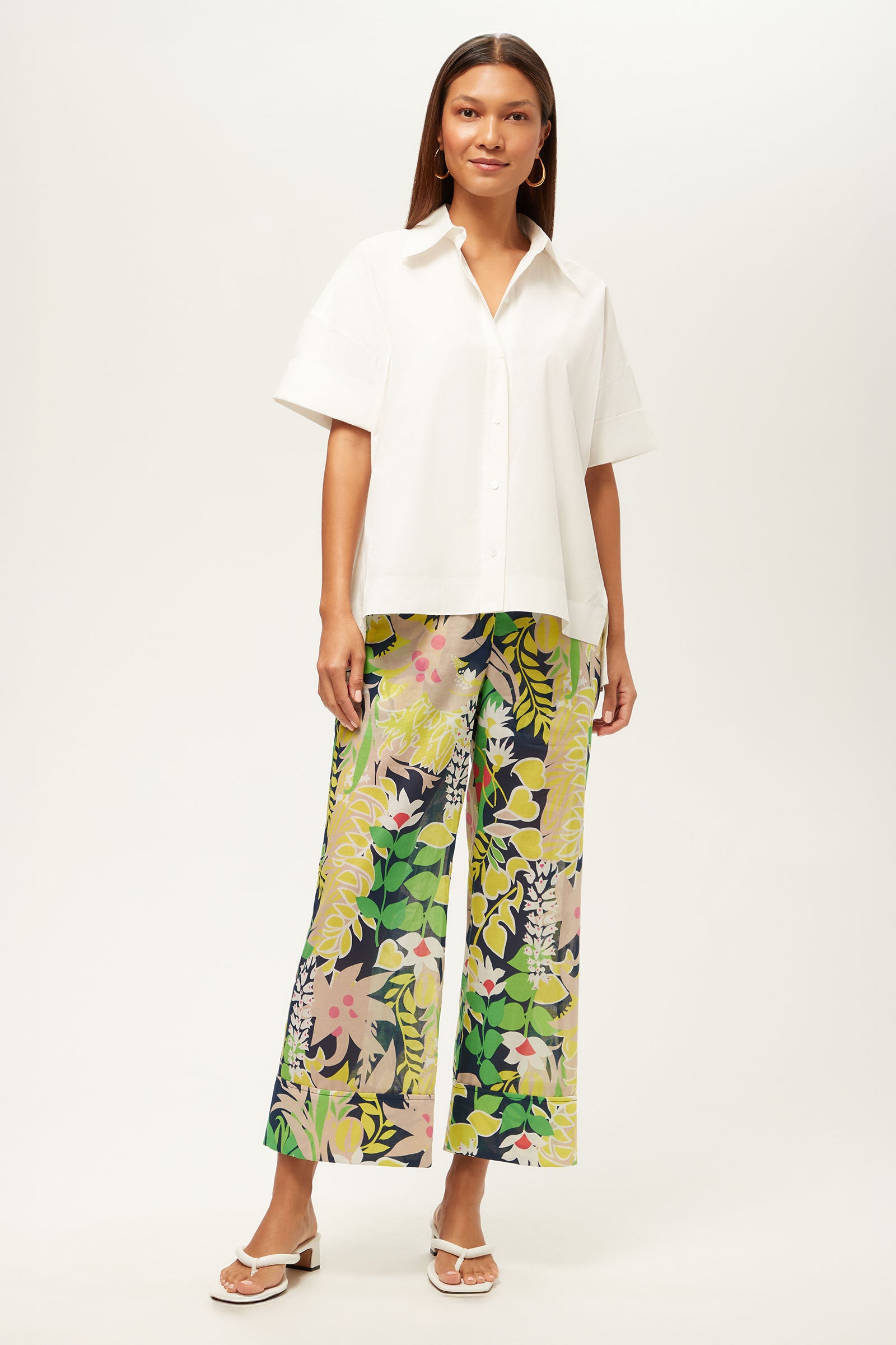 A woman stands against a plain white background wearing the Opponent Short-Sleeve Button-Up Top, colorful wide-leg floral pants in green, yellow, and pink, and white sandals.