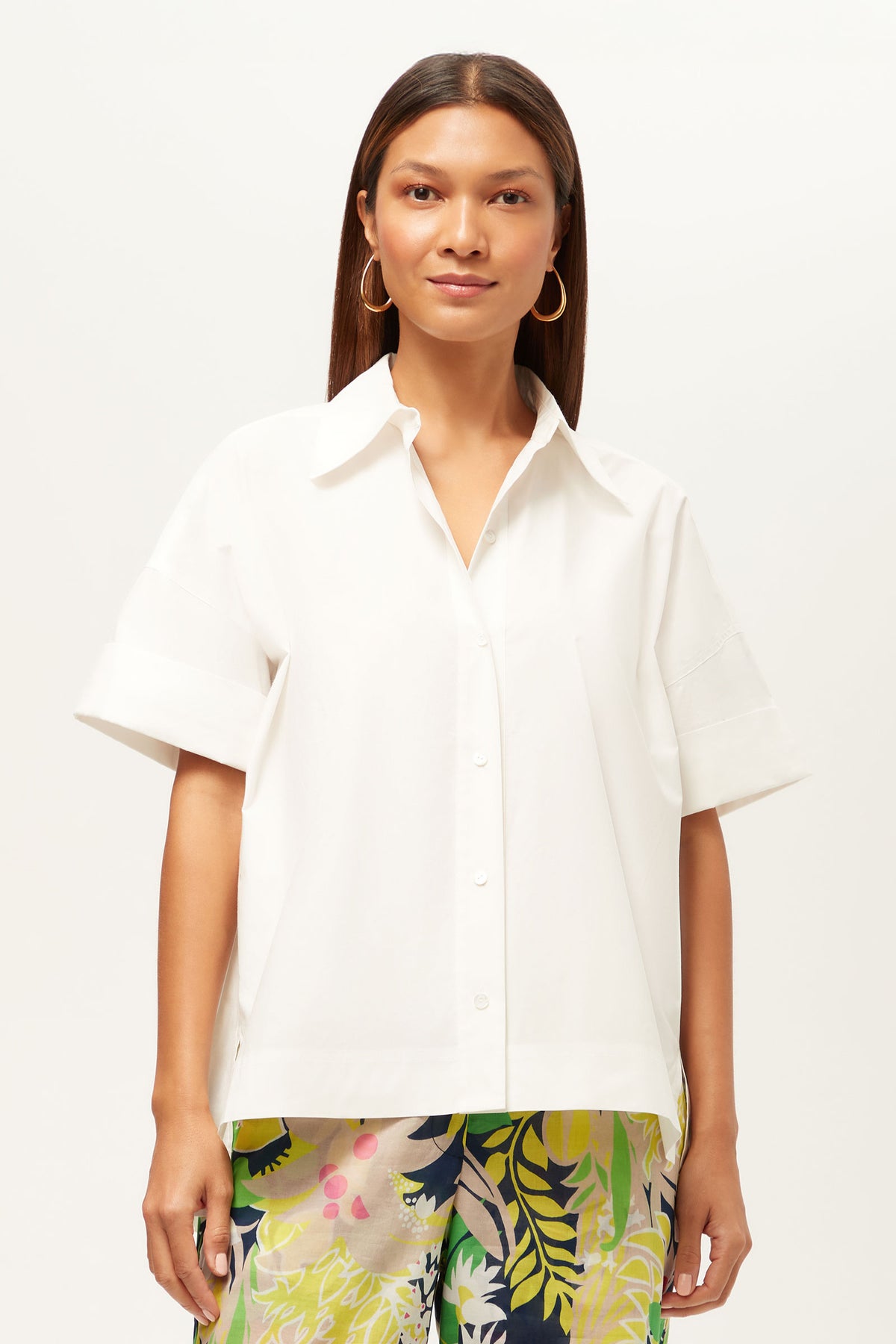 A woman with straight brown hair wears the white Opponent Short-Sleeve Button-Up Top and colorful floral pants, standing facing forward against a plain light background.