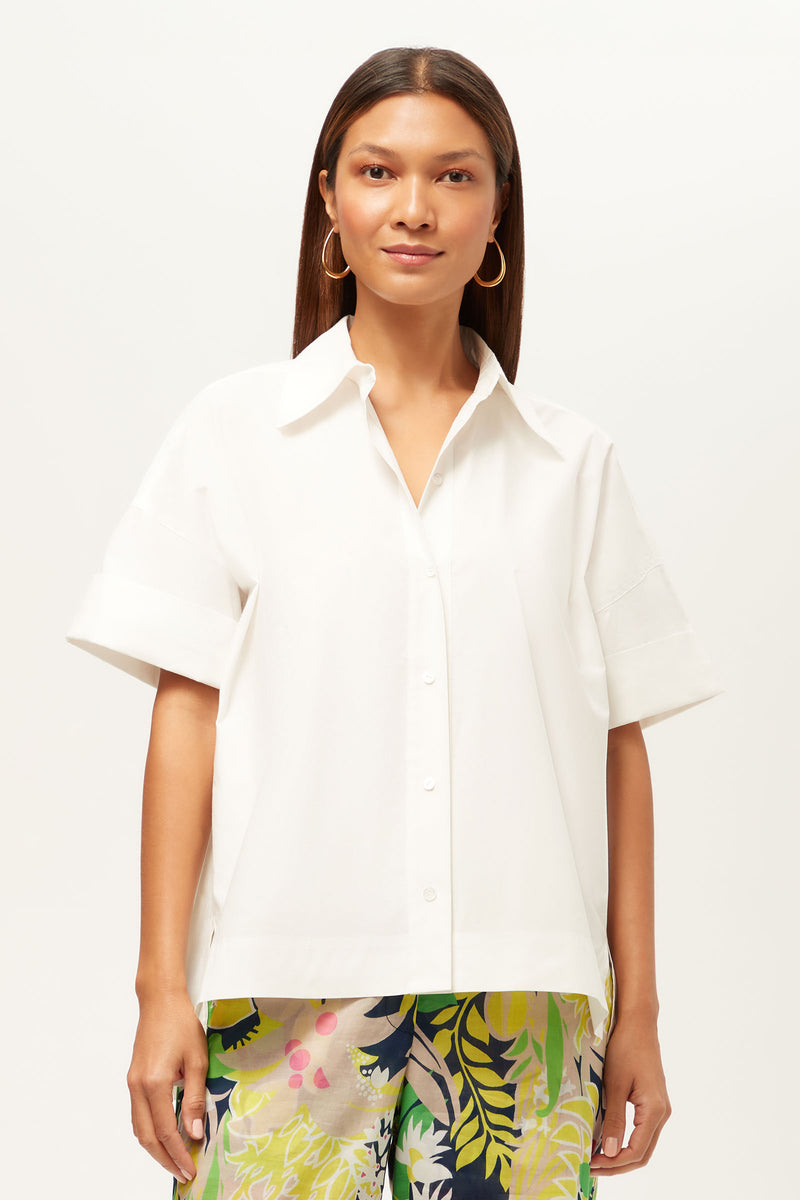 A woman with straight brown hair wears the white Opponent Short-Sleeve Button-Up Top and colorful floral pants, standing facing forward against a plain light background.