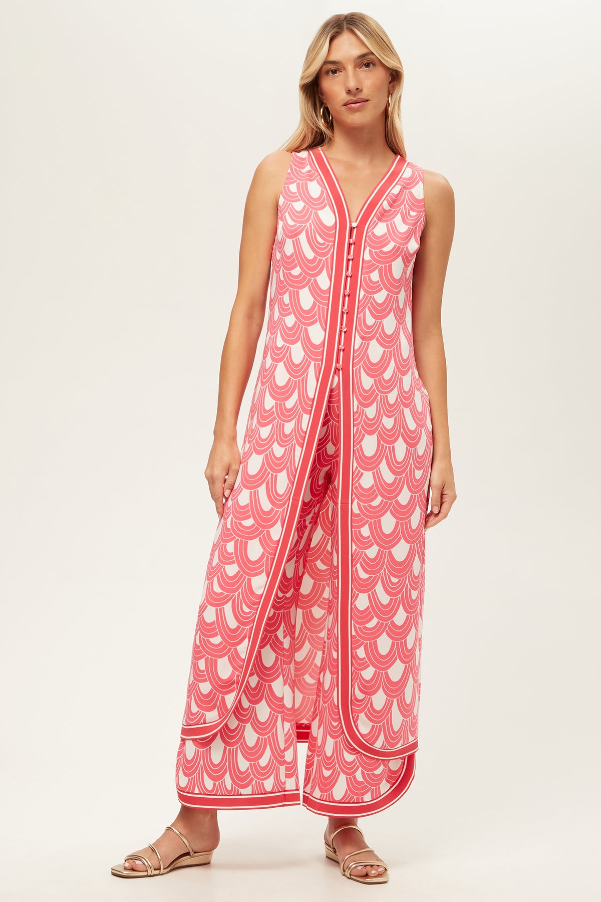 A woman in the Slice Sleeveless Maxi Top - Ace Arches in Coral Crush/Whitewash, featuring a V-neck, wide legs, and side slits, pairs it with flat sandals while standing against a plain light background.