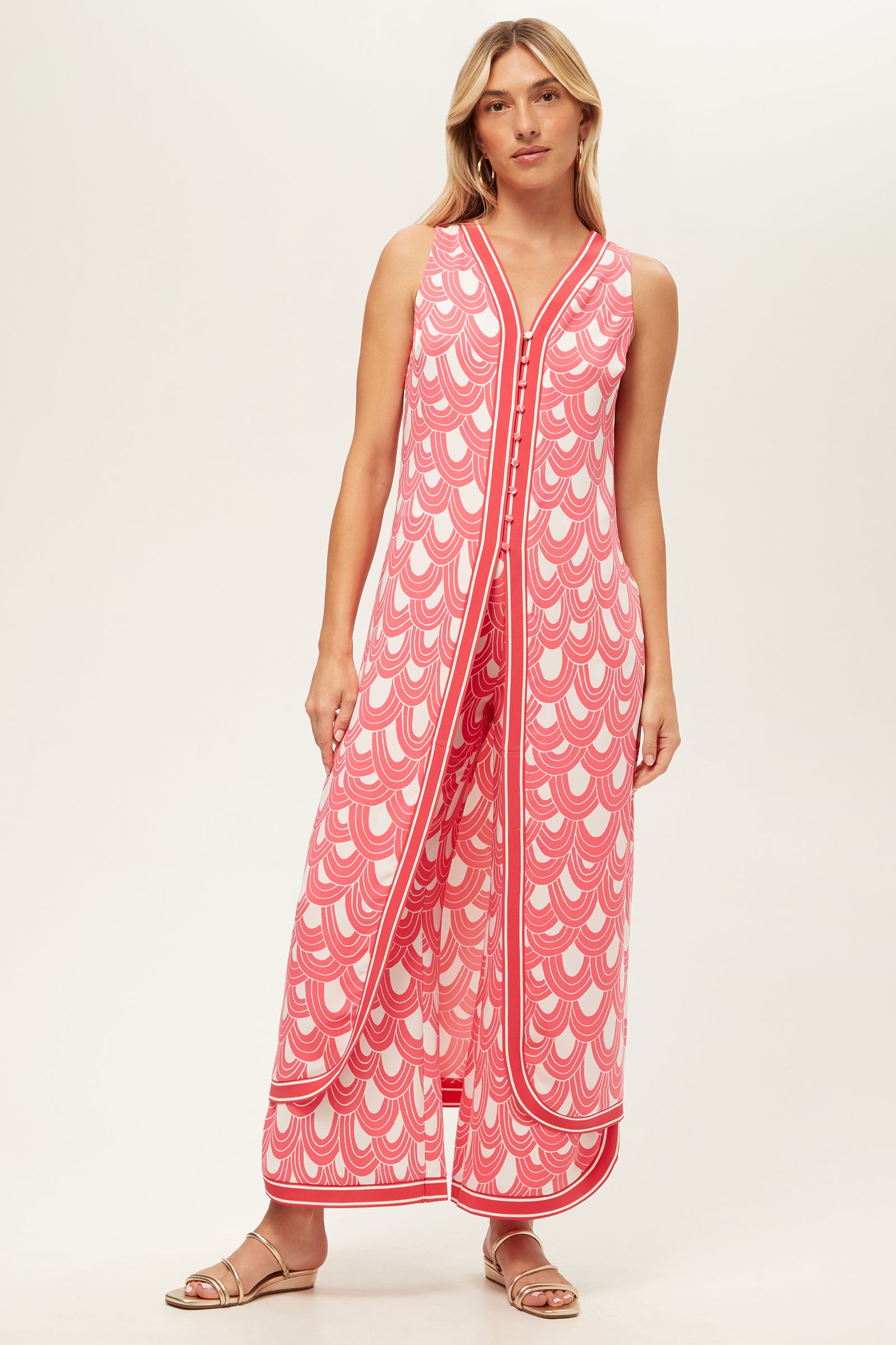 A woman with long blonde hair wears the Overhead Wide-Leg Pant - Ace Arches, a sleeveless pink and white patterned jumpsuit featuring a V-neck and wide legs. She pairs it with flat sandals against a plain light background.