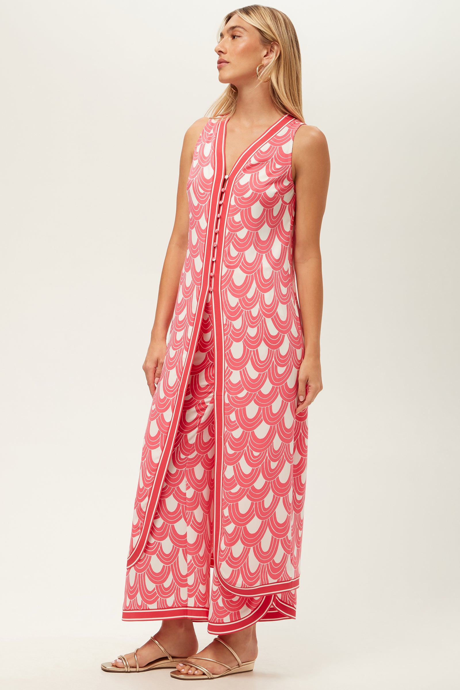 A woman wears the Slice Sleeveless Maxi Top - Ace Arches, an ankle-length pink and white dress with scalloped patterns, front buttons, and side slits. She has straight blonde hair and flat sandals.