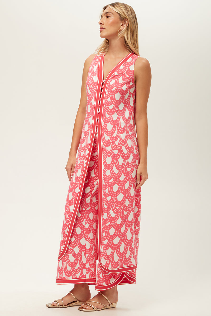 A woman wears the Slice Sleeveless Maxi Top - Ace Arches, an ankle-length pink and white dress with scalloped patterns, front buttons, and side slits. She has straight blonde hair and flat sandals.