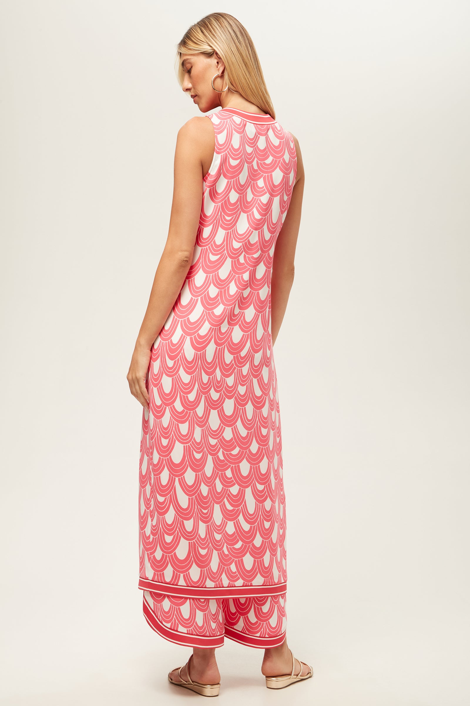 A woman with long blonde hair wears the Slice Sleeveless Maxi Top in Ace Arches, featuring a pink and white scallop pattern, paired with flat sandals. She stands facing away against a plain light background.