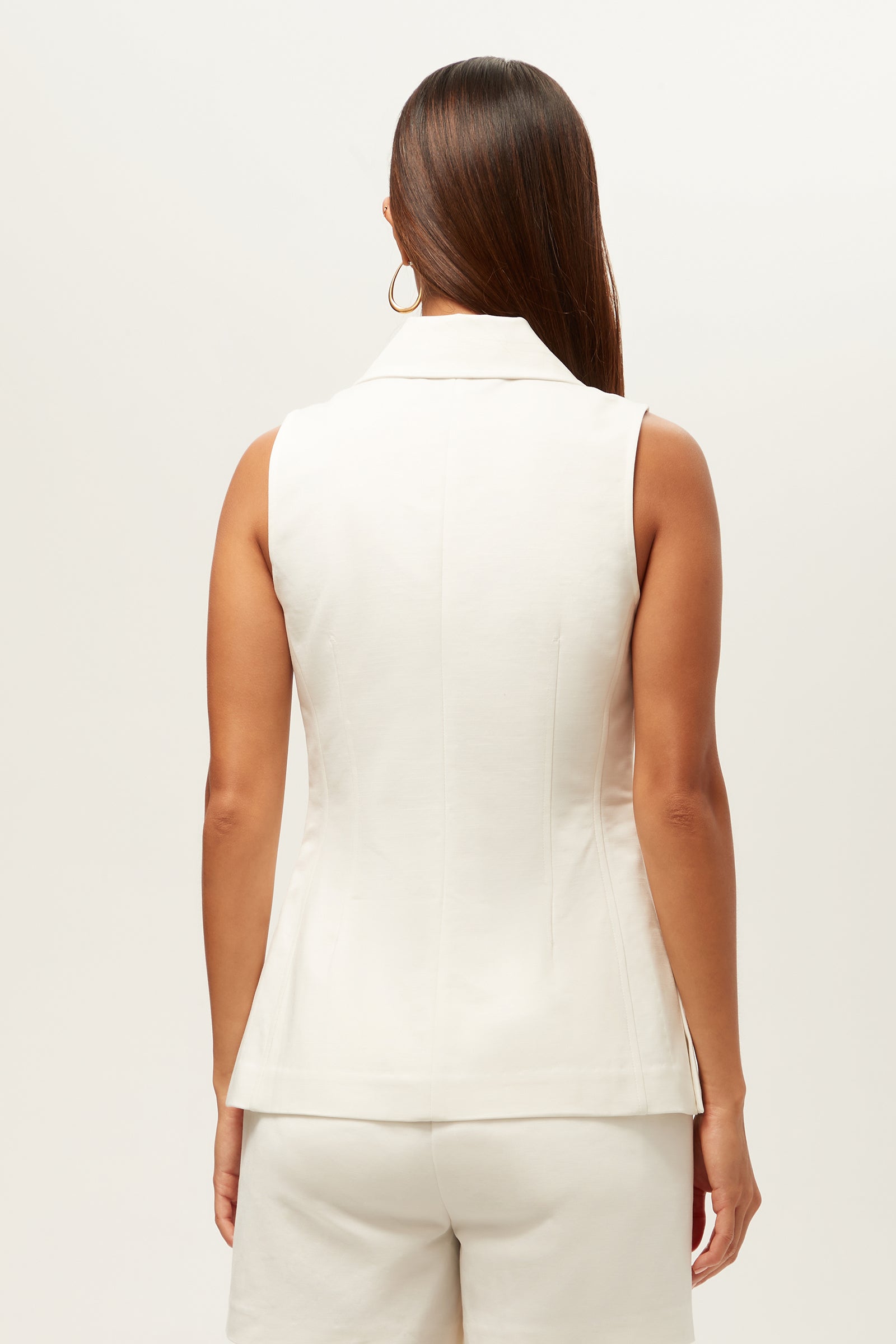 A woman with long brown hair, wearing hoop earrings, is shown from the back in the Follow Through Sleeveless Top - Solid Suiting and matching white shorts against a plain, light background.