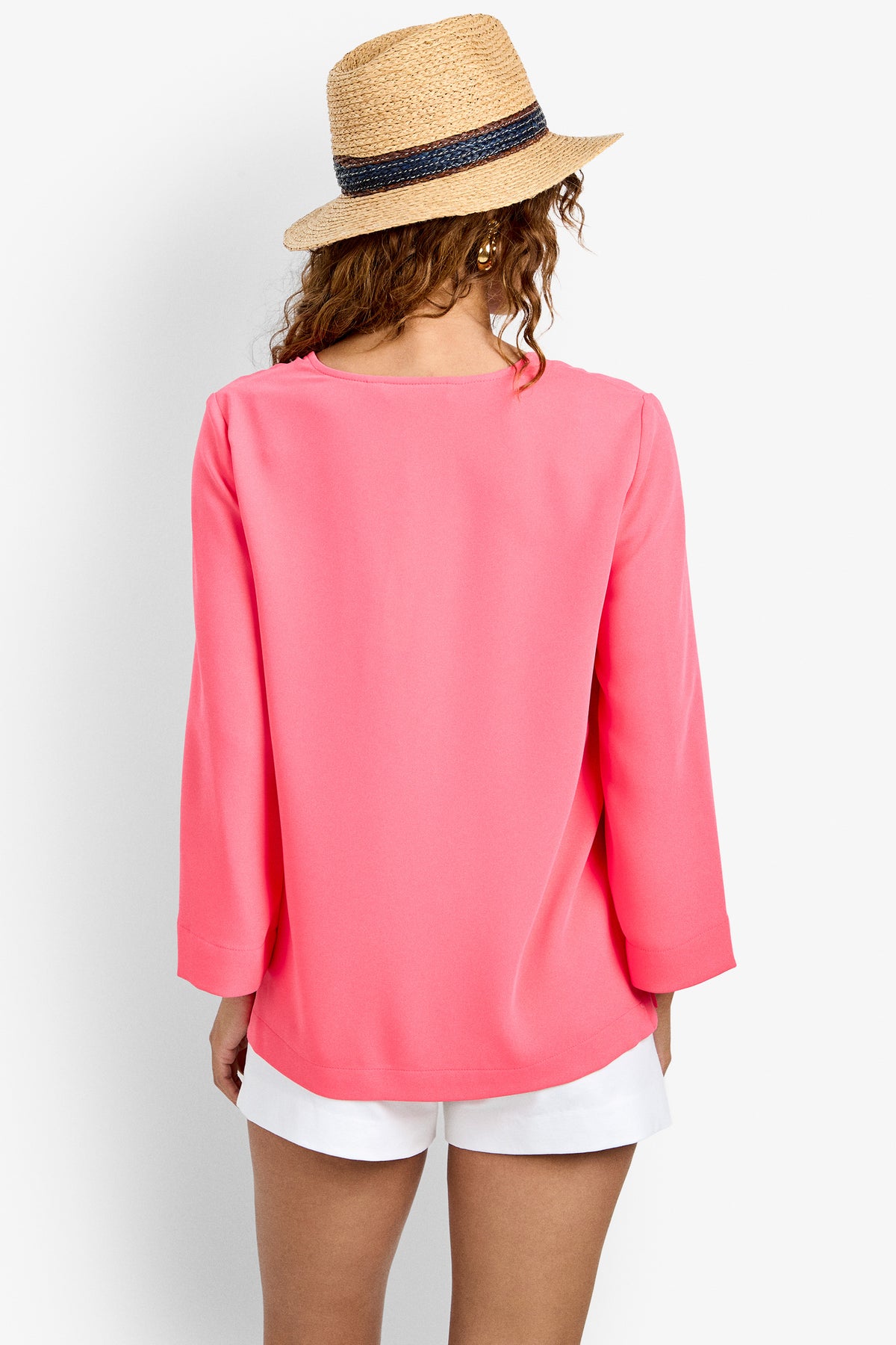 A woman stands with her back to the camera against a plain white background, wearing a straw hat, white shorts, and the bright pink Catamaran Top with long sleeves.