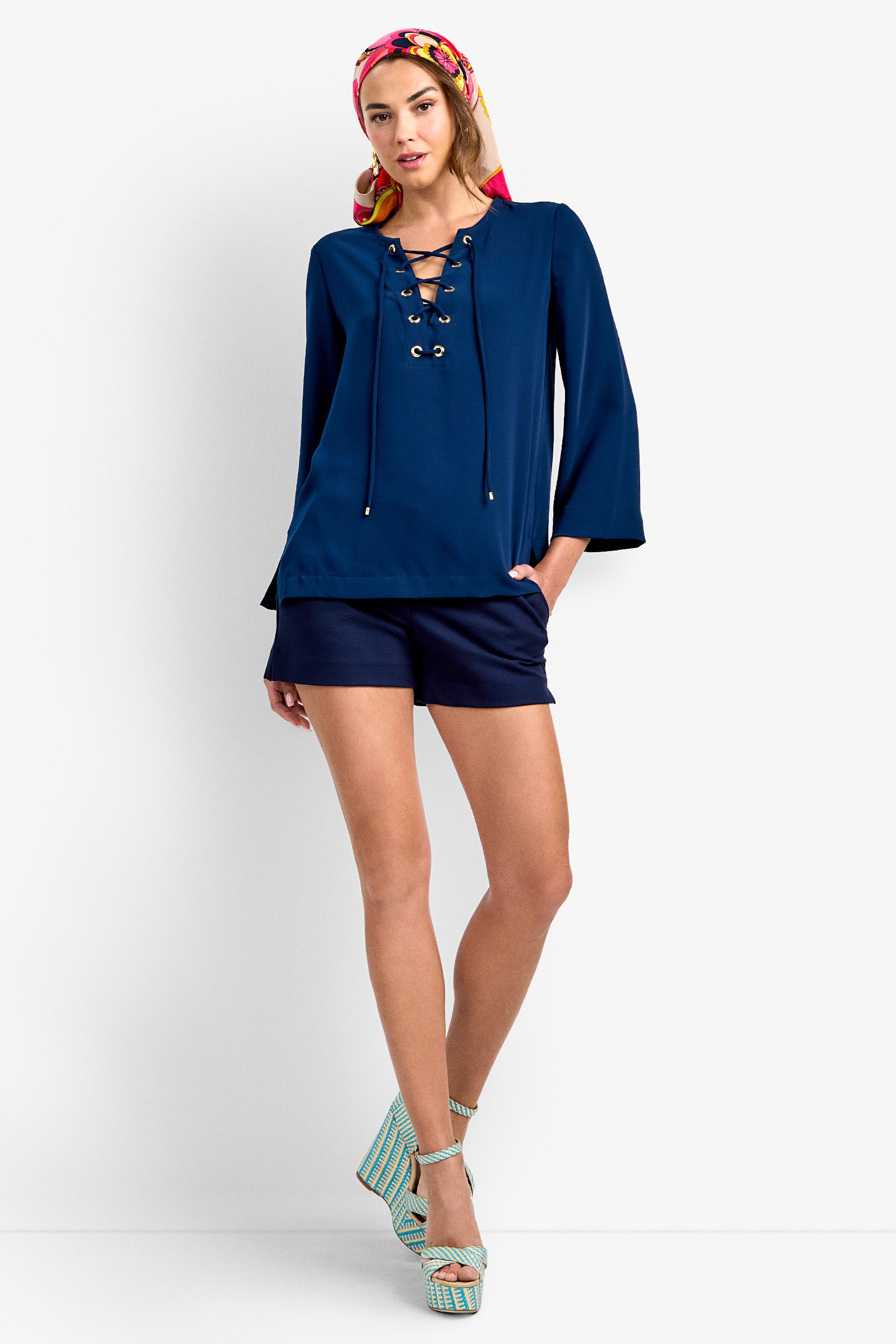 A woman in the Catamaran Top in INK, navy shorts, striped platform sandals, and a colorful headscarf stands against a white background with one hand in her pocket.