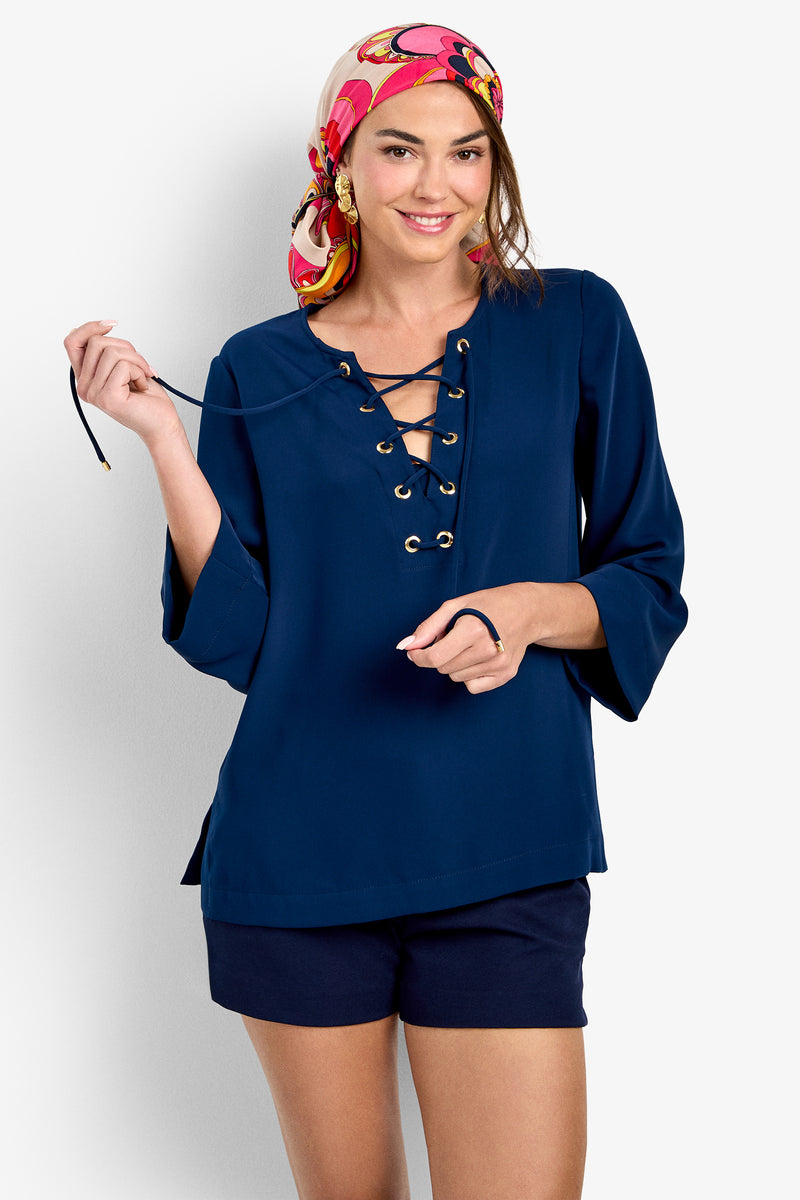 A woman stands smiling against a white background, wearing a colorful headscarf, dark shorts, and the Catamaran Top—a navy blue lace-up blouse with grommet details.