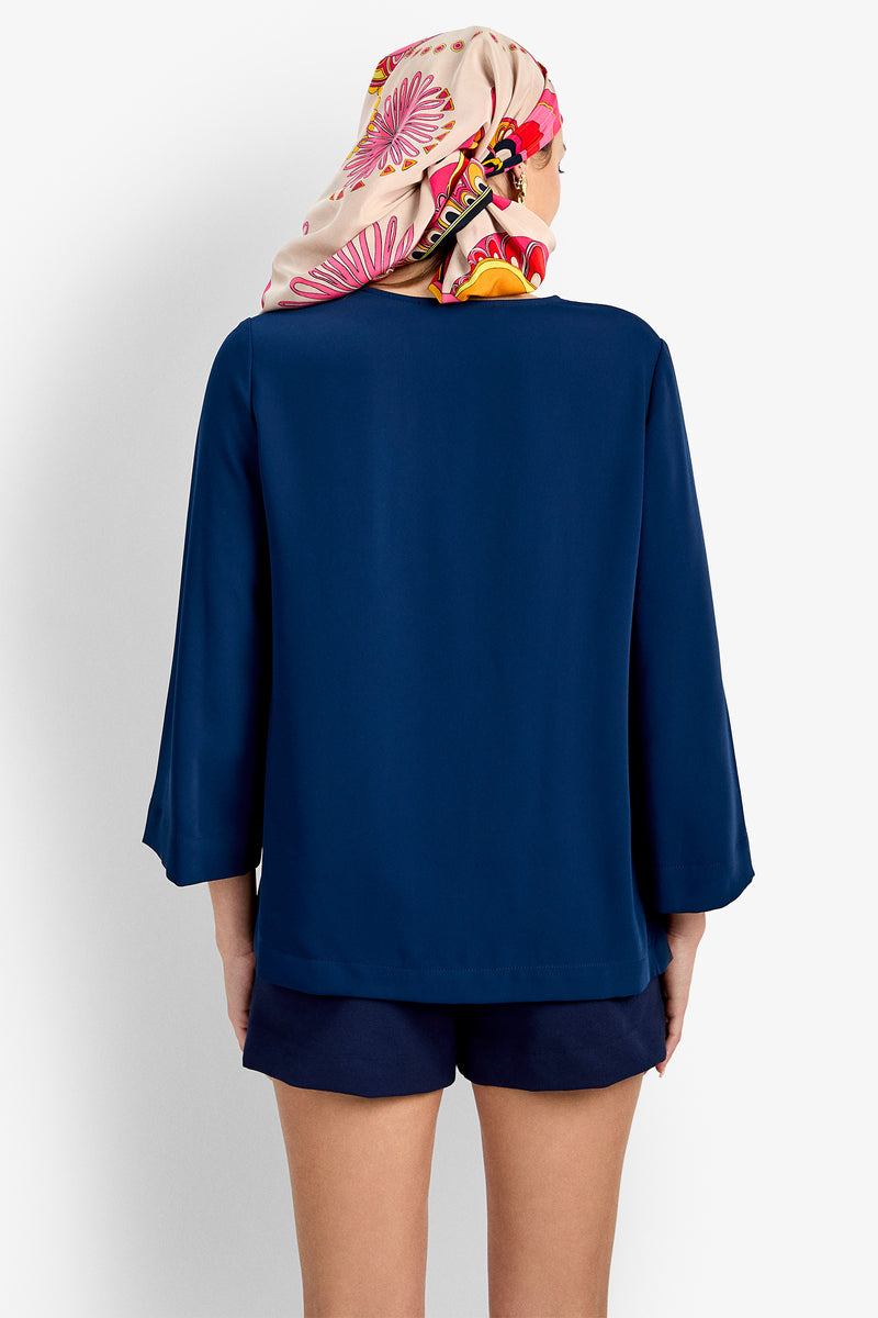 A person facing away from the camera wears a navy blue Catamaran Top, matching navy shorts, and a colorful scarf with pink and orange patterns covering their hair.
