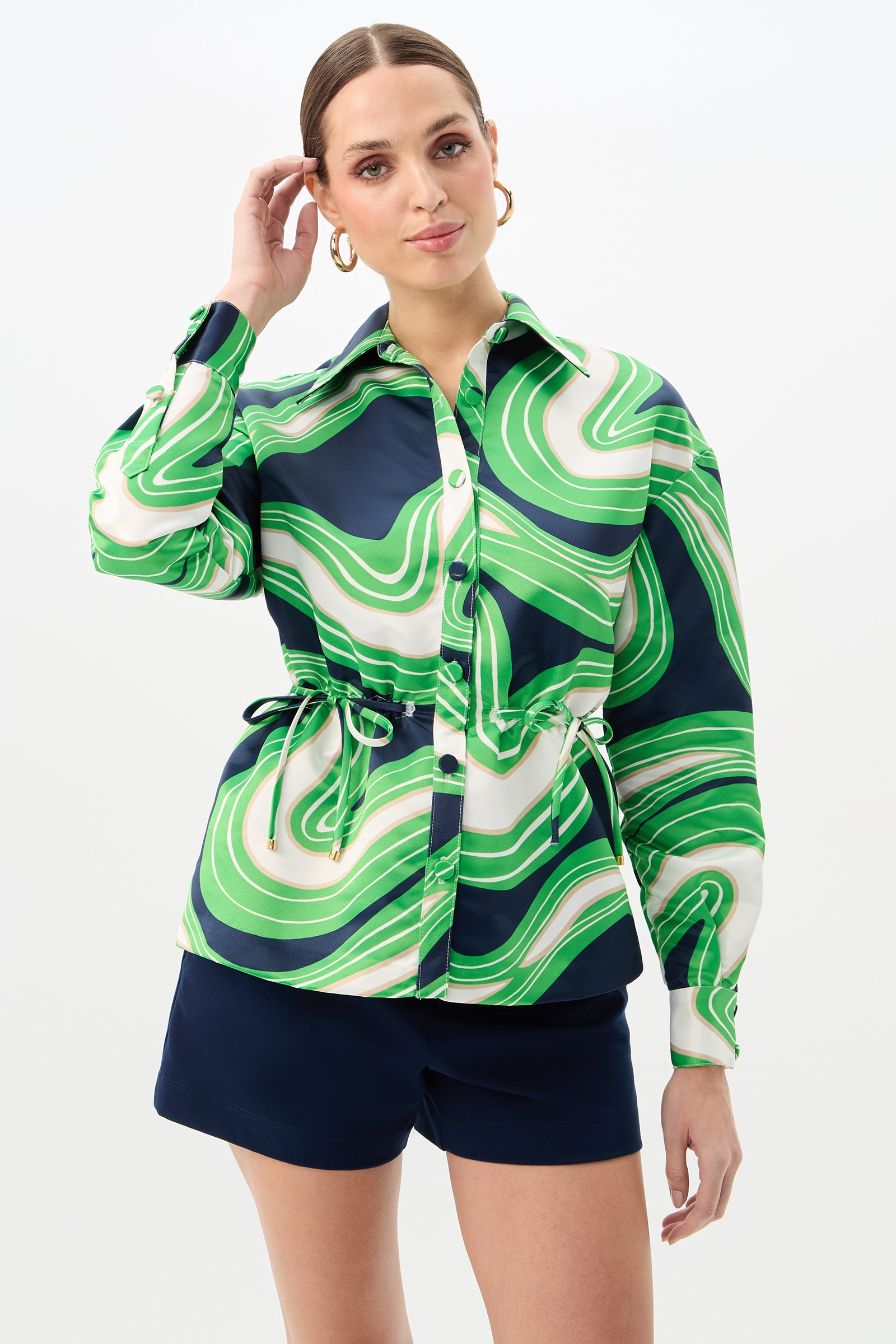 A woman wearing the Ellena Drawstring Waist Jacket in Serena Satin stands against a plain white background, looking at the camera and touching her hair. She accessorizes with gold hoop earrings and navy blue shorts.