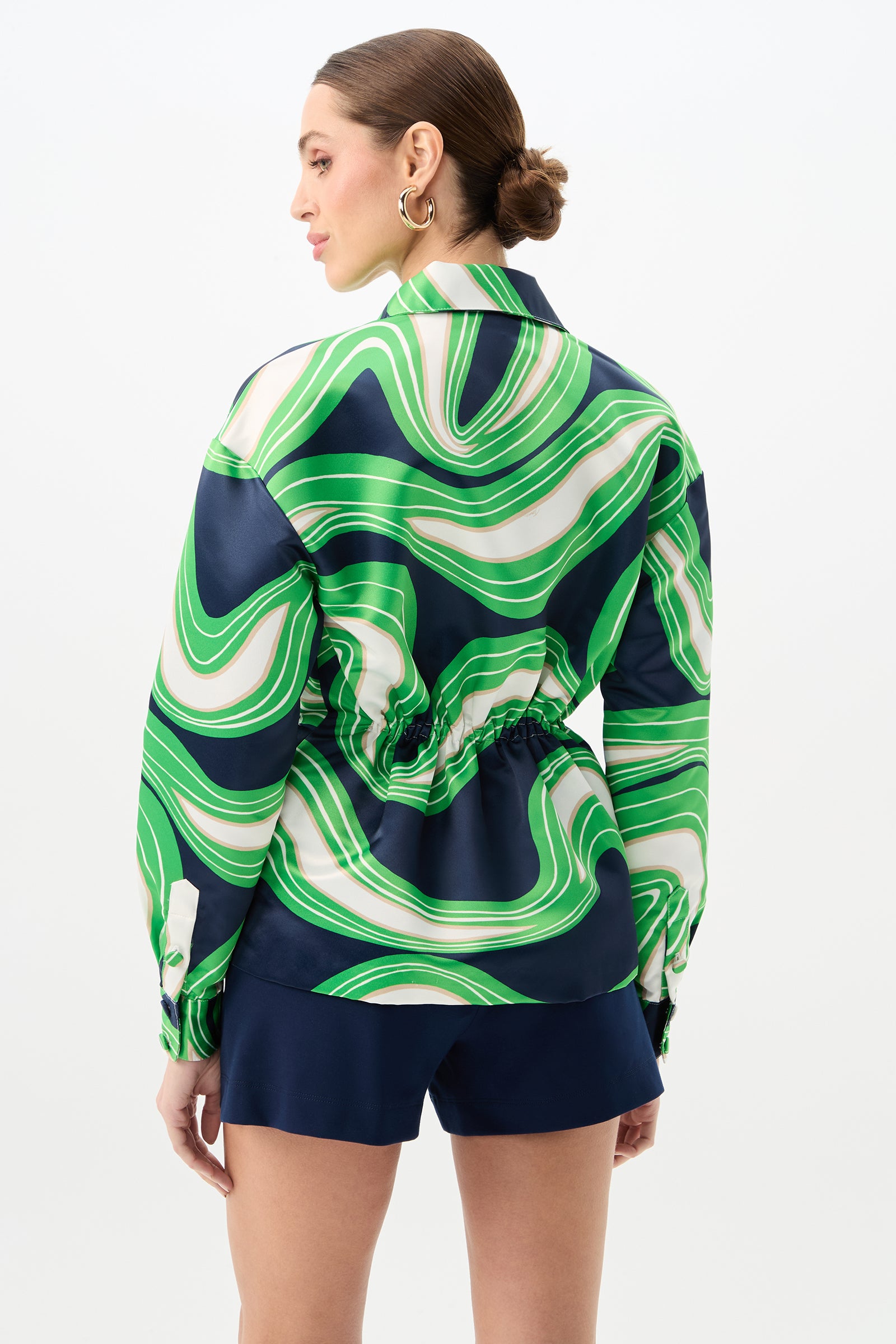 A woman faces away wearing the Ellena Drawstring Waist Jacket in Serena Satin, featuring a green, white, and navy swirl pattern with a cinched waist. She pairs it with navy shorts, gold hoop earrings, and has her hair in a neat bun.