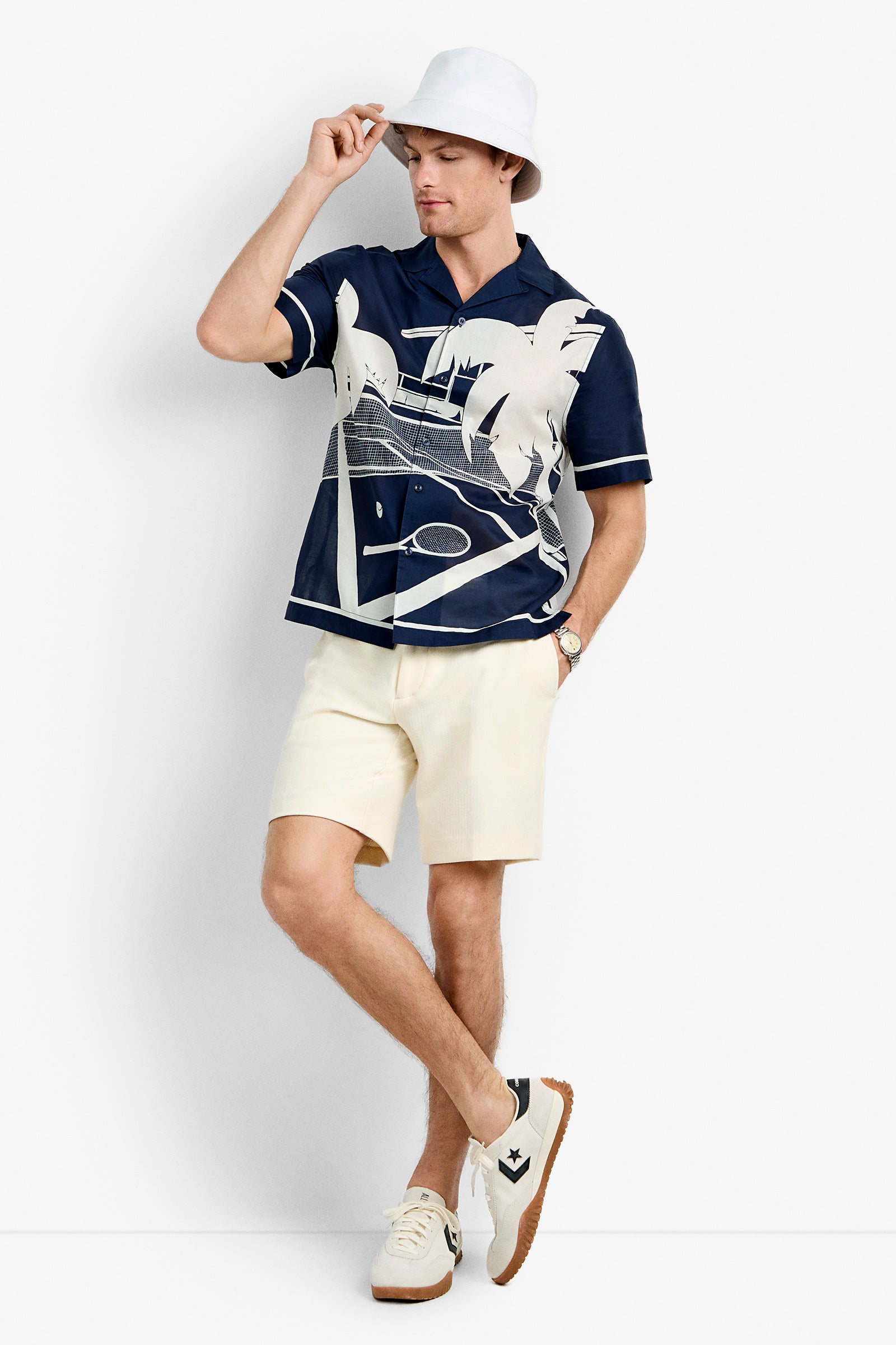 A man wears the Josue Short-Sleeve Shirt in Ink/Whitewash tennis print, a white bucket hat, cream shorts, white socks, and sneakers. He stands against a white background, touching his hat’s brim with one hand and crossing his legs.