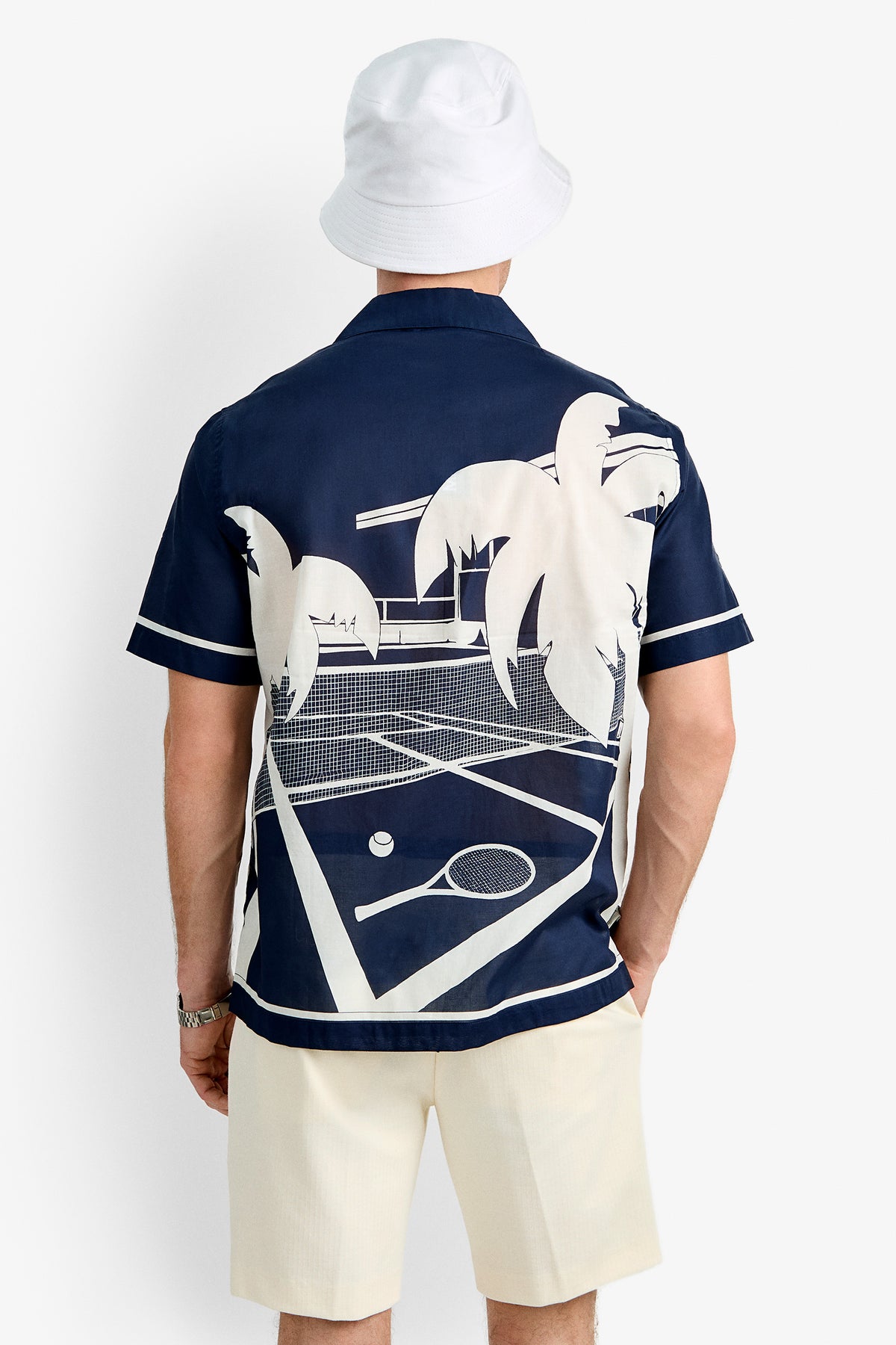 A man stands with his back to the camera, wearing a white bucket hat, cream shorts, and the Josue Short-Sleeve Shirt - Tennis Print featuring large white palm trees, tennis court, ball, and racket graphics against a white background.