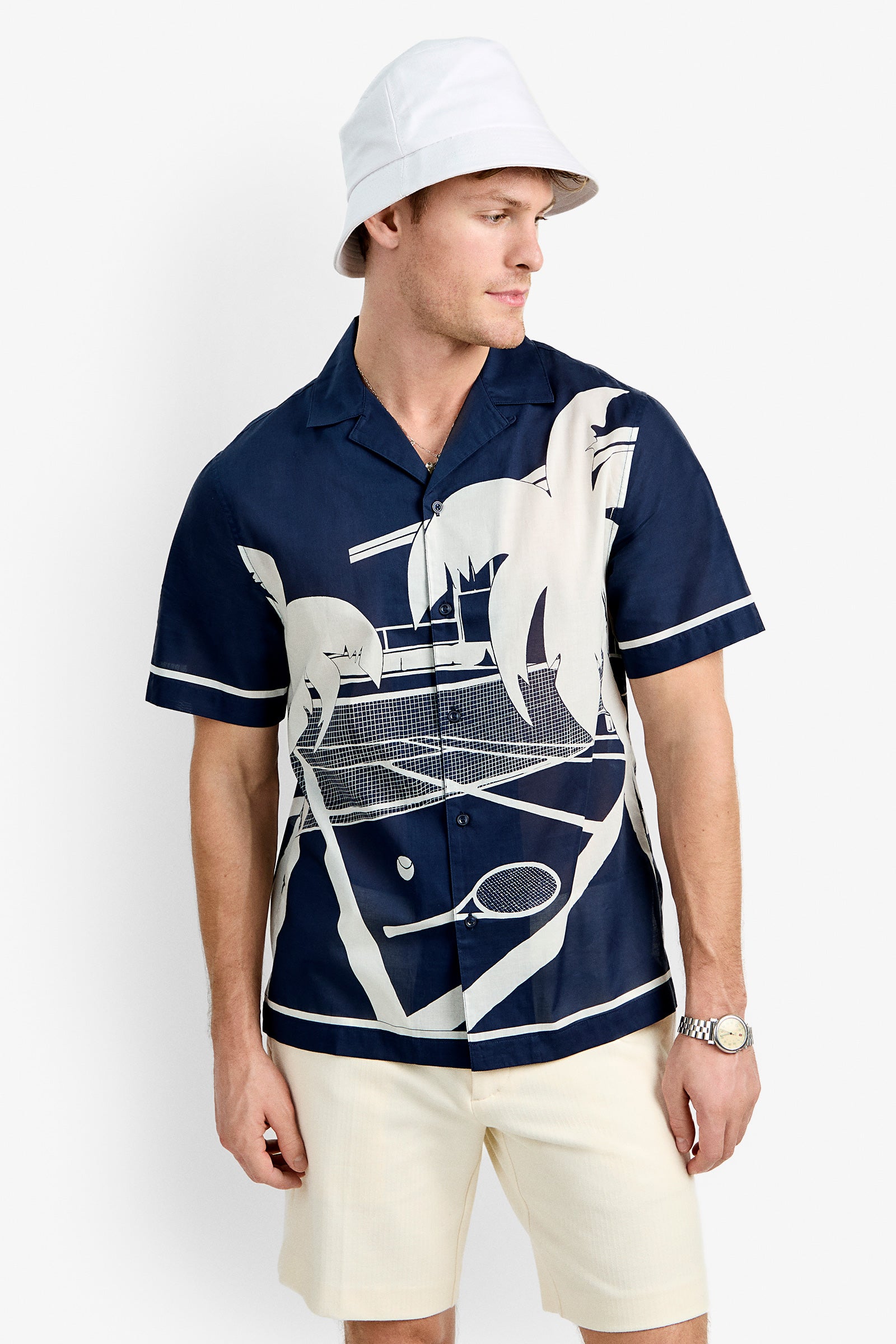 A man wearing the Josue Short-Sleeve Shirt – Tennis Print, a white bucket hat, cream shorts, and a silver wristwatch stands against a plain white background, looking to the side.