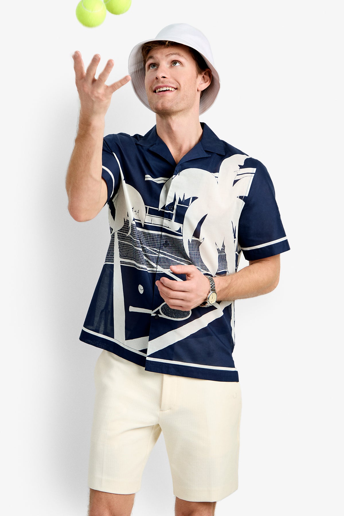 A man wearing a white bucket hat, Josue Short-Sleeve Shirt - Tennis Print, and cream shorts juggles two yellow tennis balls against a plain white background.