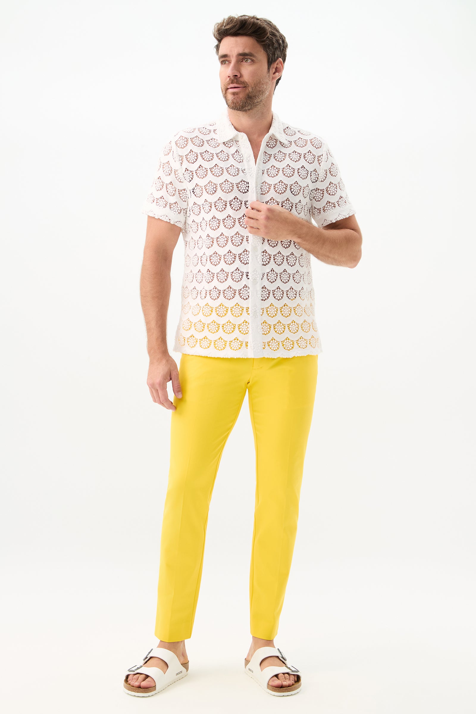 A man with short brown hair and a trimmed beard stands against a white background wearing a white lace shirt, the Clyde Slim Trouser - Sunset Stretch (bright yellow pants), and white sandals, with one hand touching his shirt.