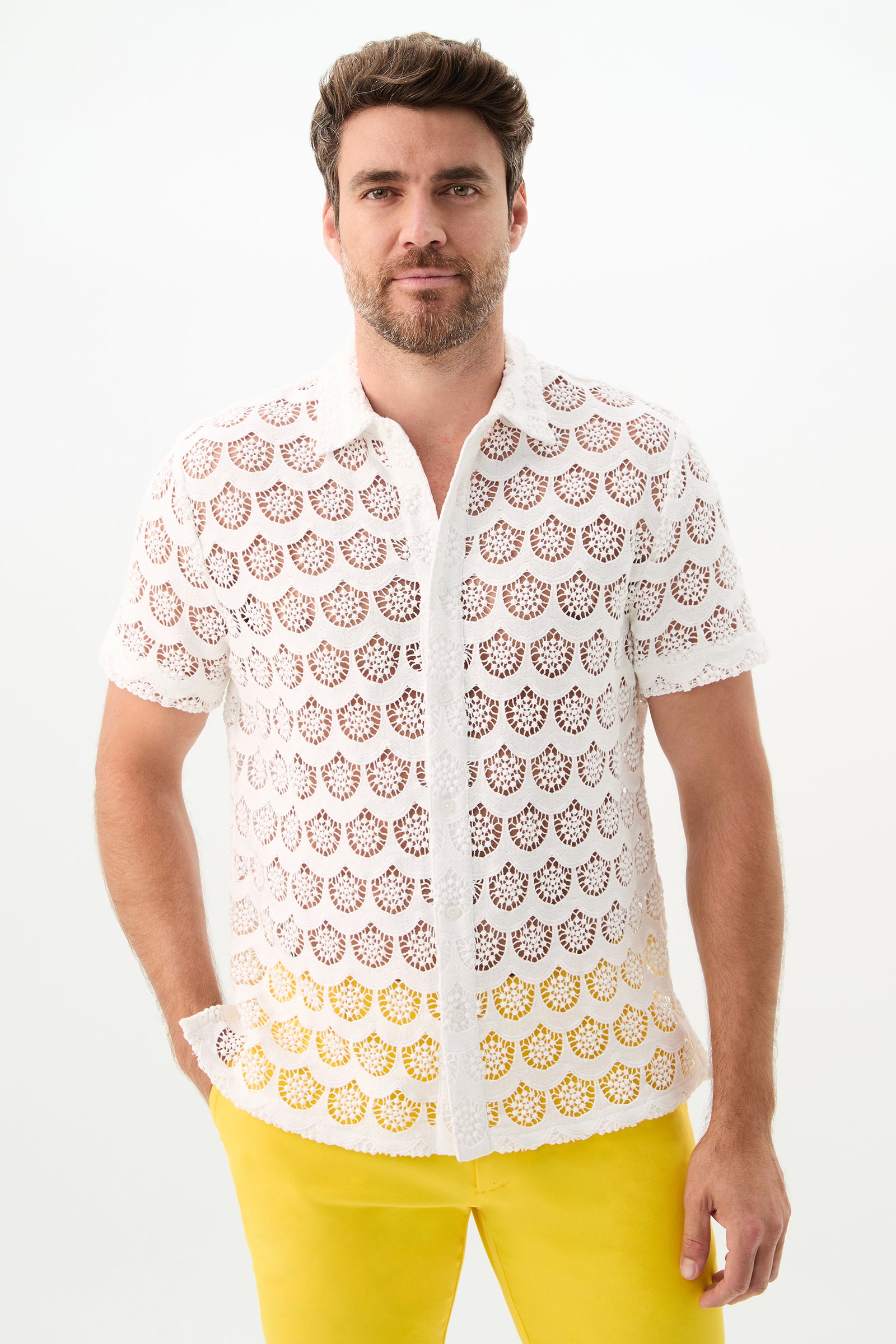 A man with short brown hair and a beard wears the Branson Short-Sleeve Shirt - Crochet Lace in white, paired with bright yellow pants, standing against a plain white background with his hands in his pockets.