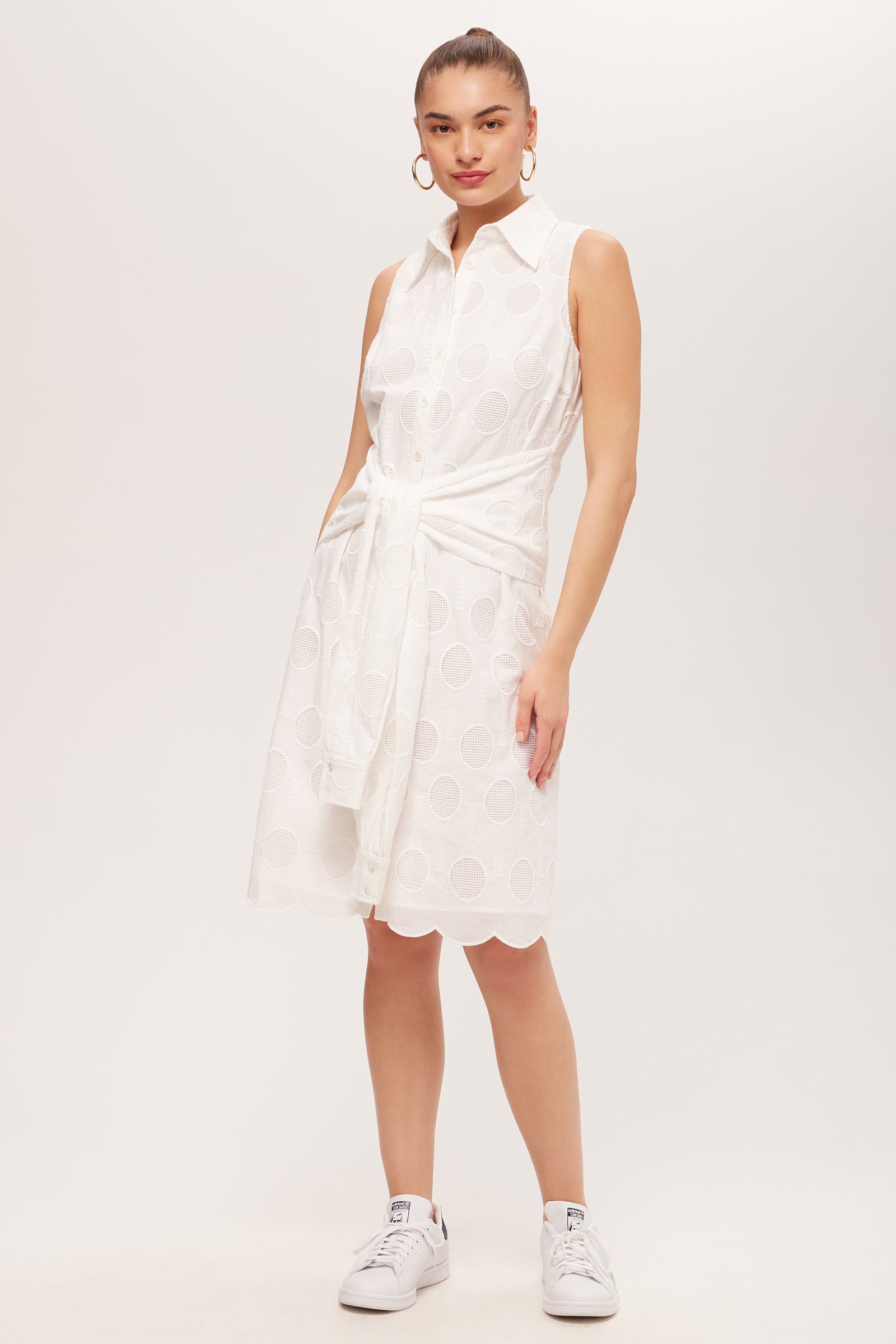 A woman stands against a plain background wearing the Rally Sleeveless Shirt Dress - Tennis Eyelet in Whitewash, featuring a tied waist, scalloped hem, and circular patterns. She pairs it with white sneakers and large hoop earrings.