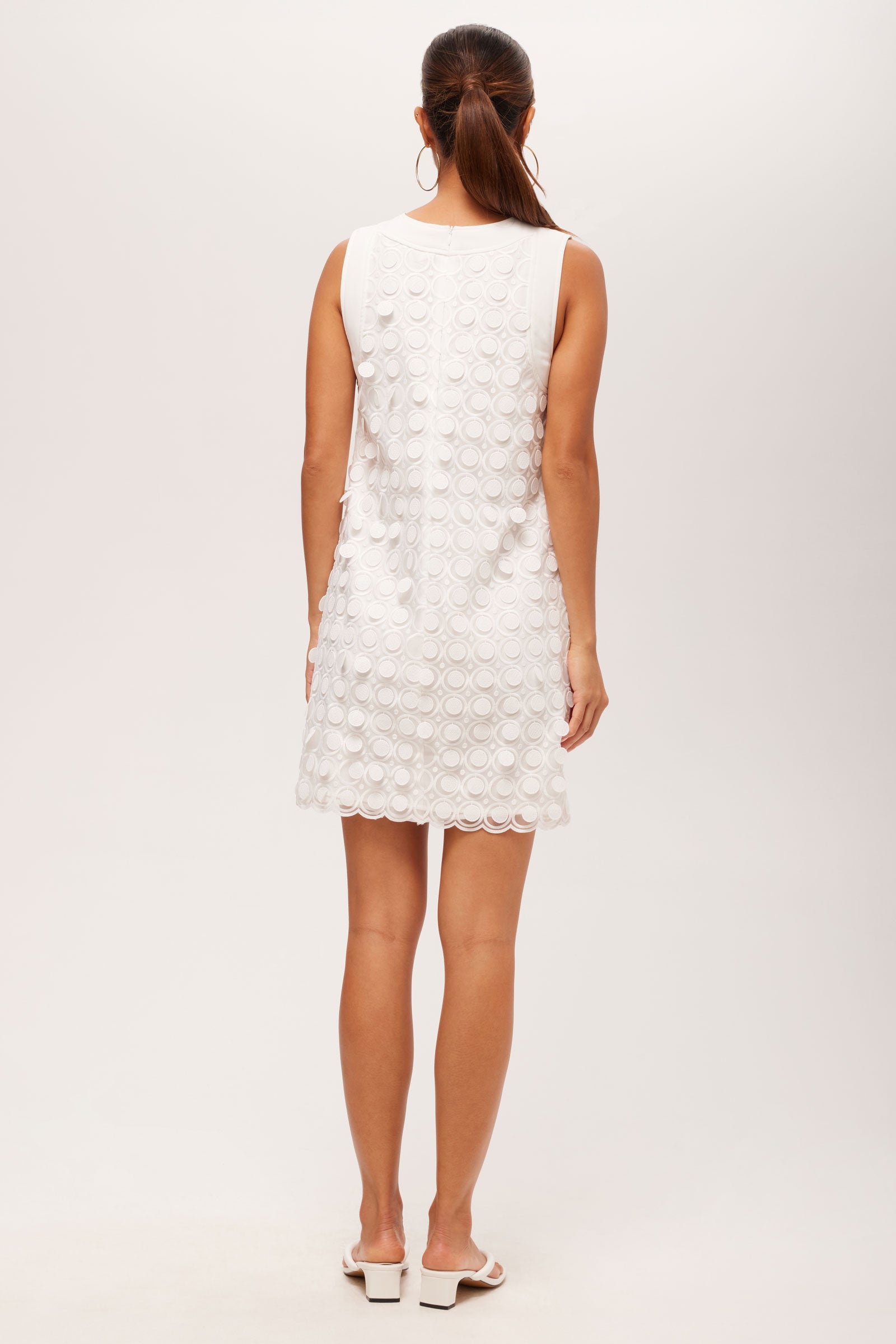 A woman faces away, wearing the Bev 2 Sleeveless Dress in Agassi Embroidery—a white textured mini dress—with white slide sandals and her hair in a ponytail, set against a plain white background.
