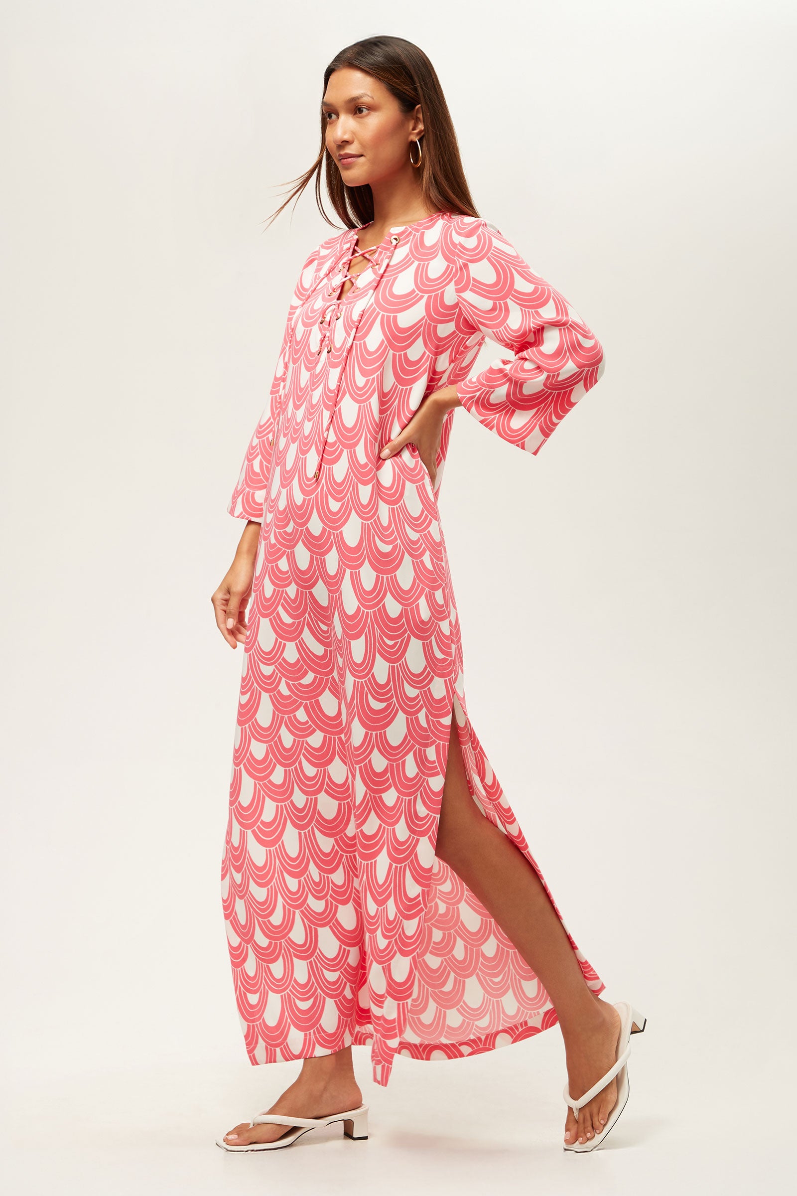 A woman models the Catamaran 3/4-Sleeve Maxi Dress, featuring a loose fit, pink and white scalloped print, side slit, and lace-up neckline. She pairs it with white sandals and gold hoop earrings against a plain background.
