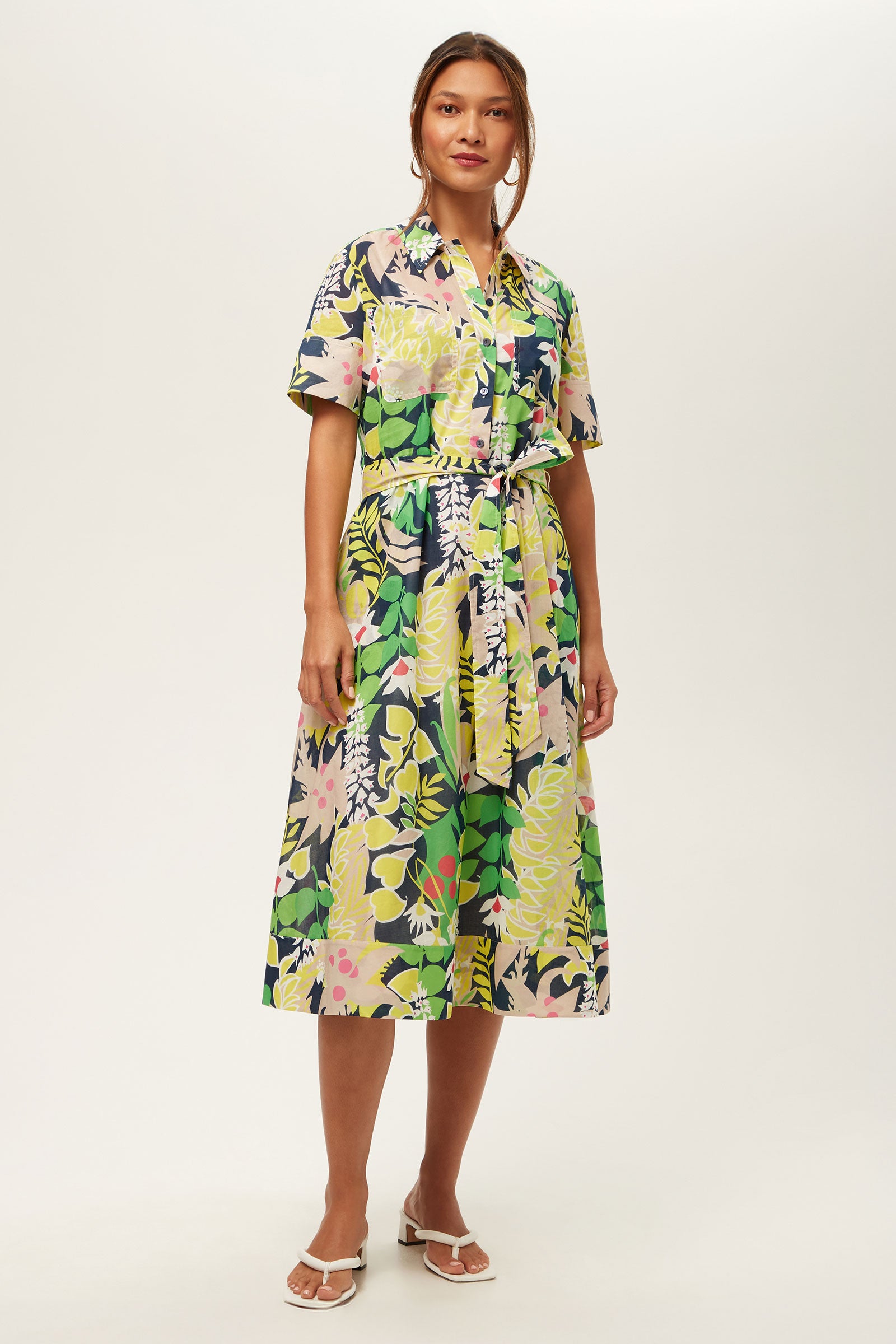 A woman wears the Misola 2 Short-Sleeve Midi Dress in Palm Springs Eternal, featuring a colorful floral print, belted waist, and midi length. She pairs it with white sandals and stands against a plain white background.