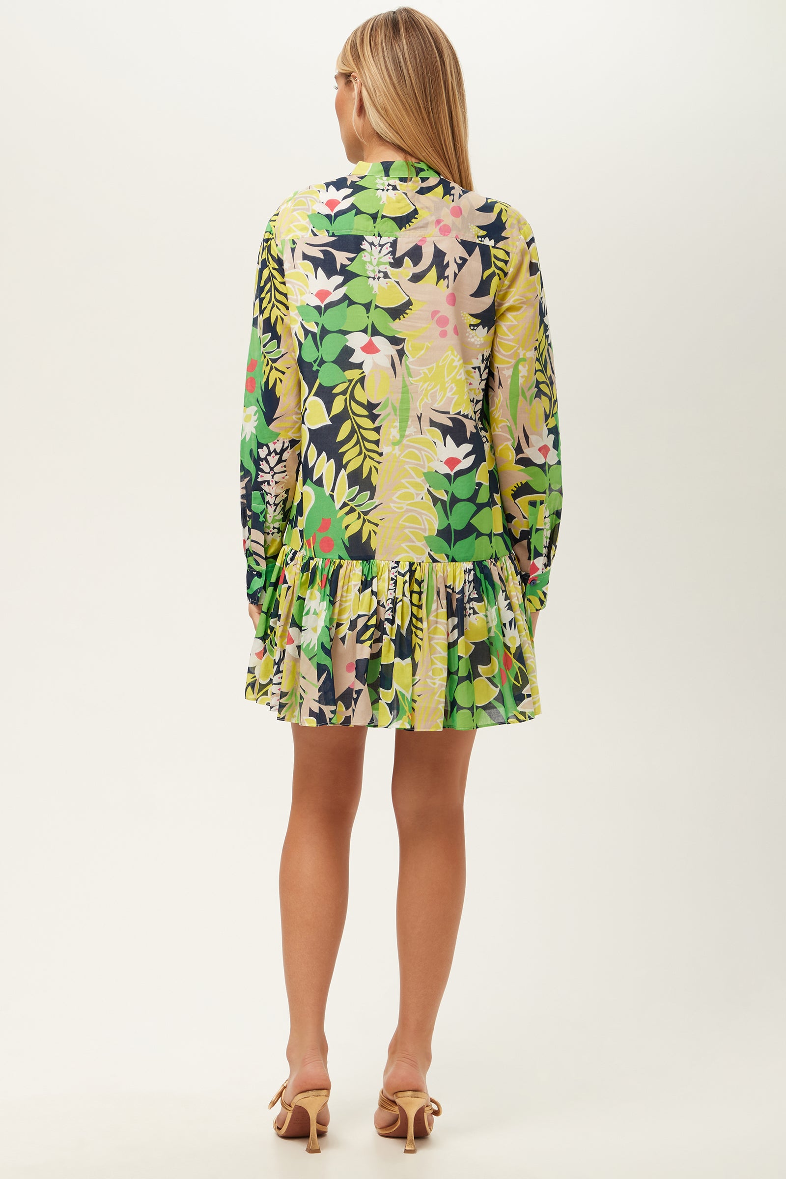 A woman with long blonde hair faces away, wearing the Championship Long-Sleeve Dress - PS Eternal featuring a colorful green, yellow, pink, and navy floral and leafy pattern, paired with light heeled sandals.