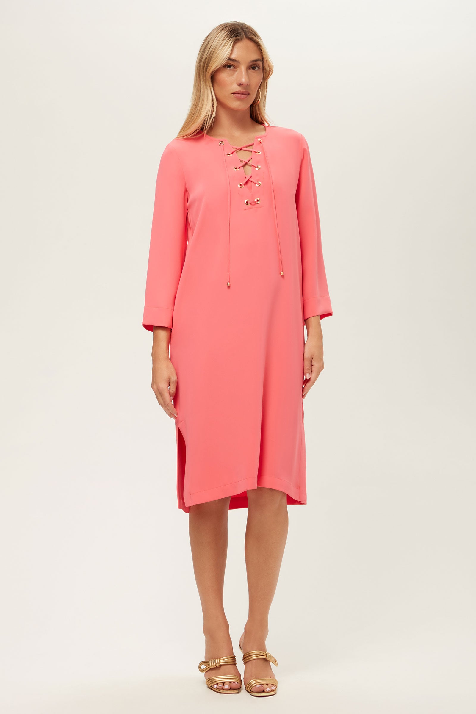A woman wears the Catamaran 3/4-Sleeve Midi Dress in coral pink with a lace-up neckline and gold-tipped ties, paired with gold sandals, standing against a plain light background.