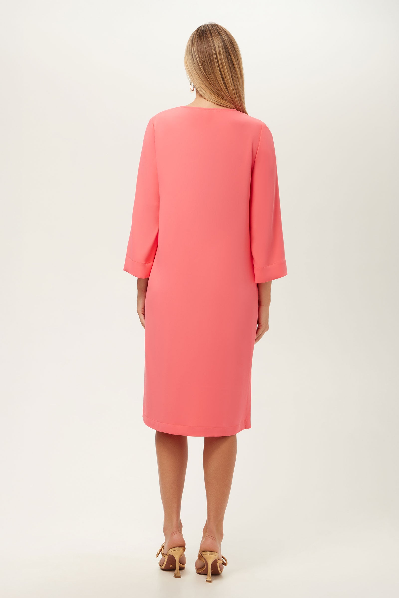 A woman faces away in a knee-length Catamaran 3/4-Sleeve Midi Dress, coral pink, with beige heeled sandals. Her straight light brown hair falls past her shoulders. The background is plain and light-colored.