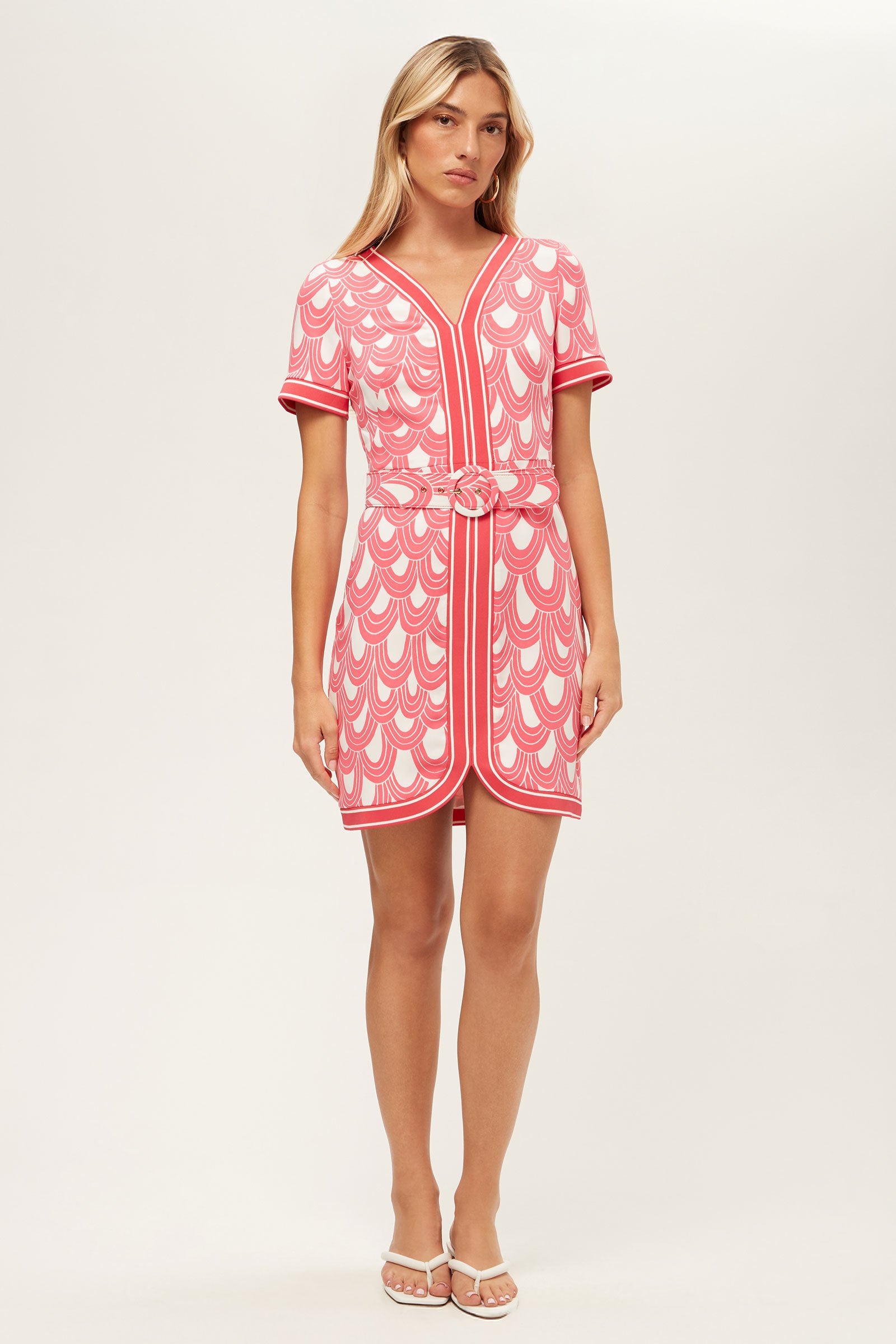A woman stands against a plain light background wearing the Motion Short-Sleeve Dress - Ace Arches in Coral Crush/Whitewash, featuring a geometric scallop pattern, belted waist, knee-length cut, and white sandals.