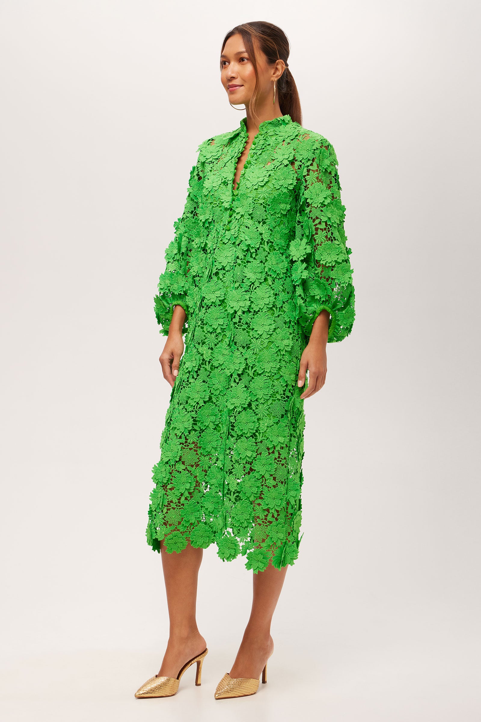 A woman in profile wears the Boheme 3/4-Sleeve Midi Dress - Love All Lace, a bright green, knee-length lace dress with floral patterns and puffed sleeves. She pairs it with gold high heels. The background is plain white.
