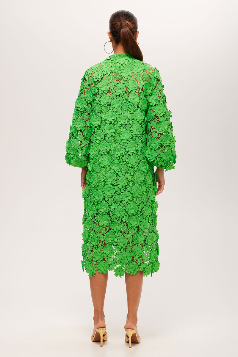 A woman faces away, wearing the Boheme 3/4-Sleeve Midi Dress - Love All Lace in vibrant green with a floral lace pattern, hoop earrings, tied-back hair, and beige heeled sandals.