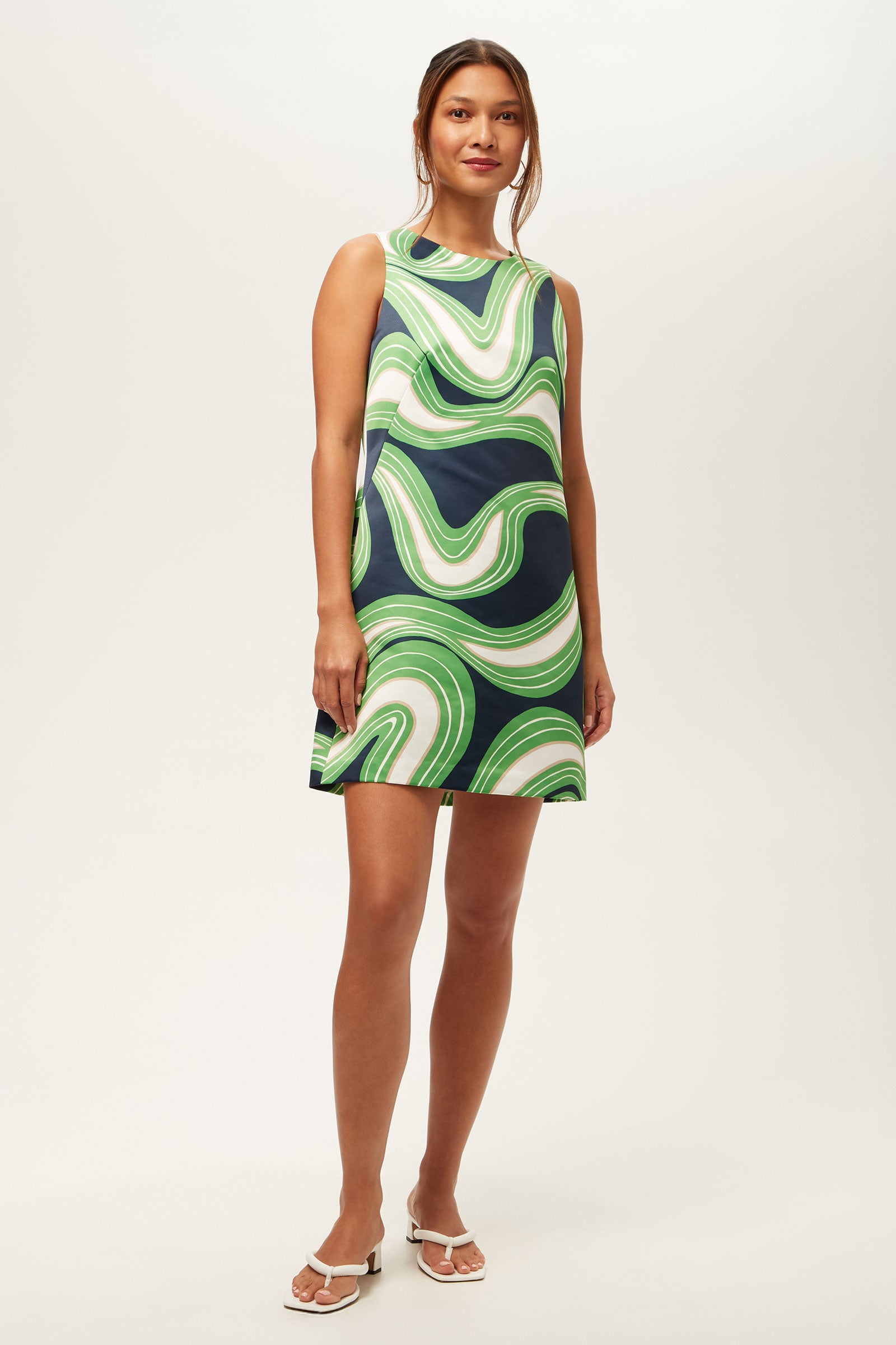 A woman stands against a plain background, wearing the Fairy Tale 2 Sleeveless Dress in Serena Satin—a knee-length dress with a bold green, navy, and white swirling pattern. Her long hair is tied back, and she pairs the look with white sandals.