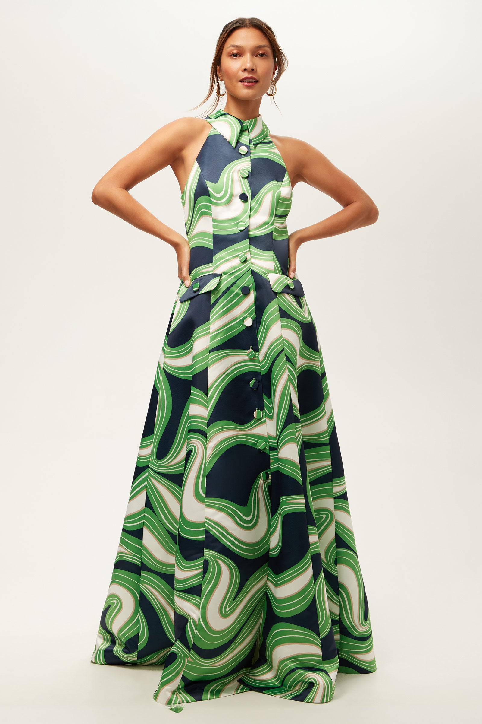 A woman stands with hands on hips, wearing the Official Sleeveless Maxi Dress - Serena Satin, featuring a bold swirling green, white, and navy pattern with button details down the front and a fitted bodice.