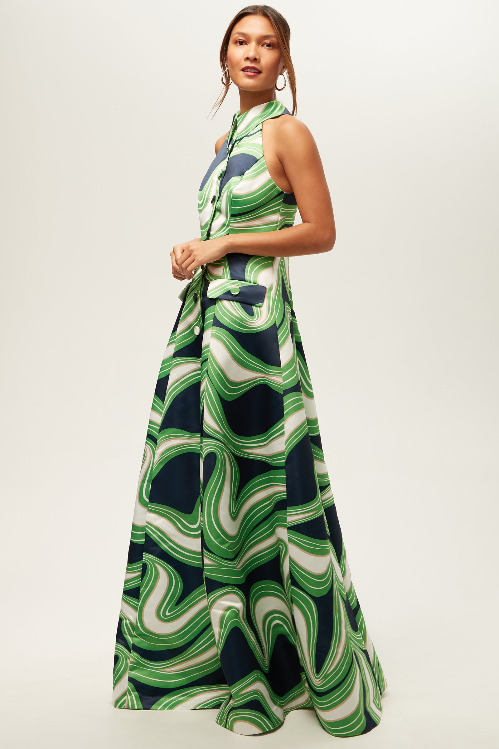 Woman wearing the Official Sleeveless Maxi Dress - Serena Satin, a floor-length dress with a bold green, white, and navy swirl pattern, standing against a plain light background.