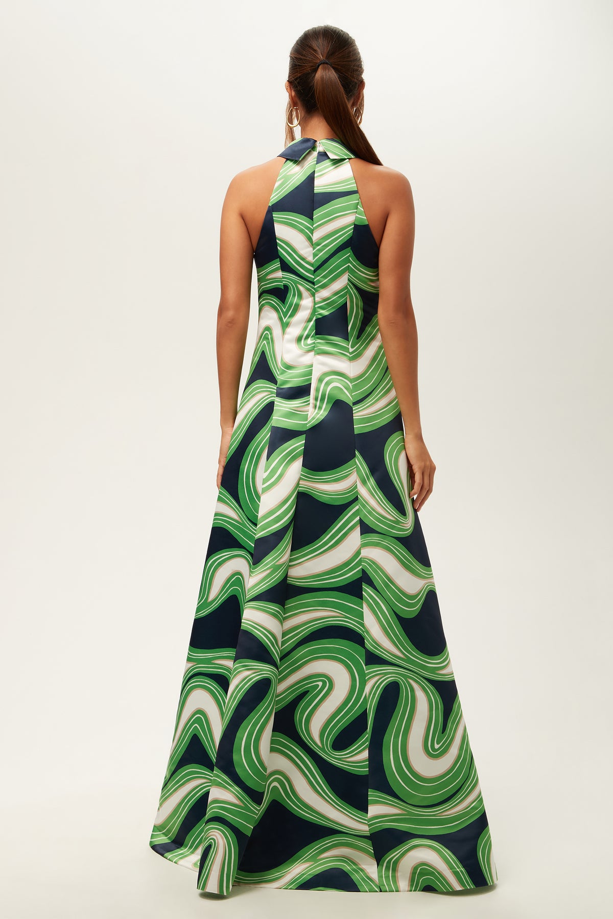 A woman stands facing away in the Official Sleeveless Maxi Dress - Serena Satin, a long halter-neck dress with a bold green and white swirling pattern accented by navy blue. Her hair is pulled back in a low ponytail.