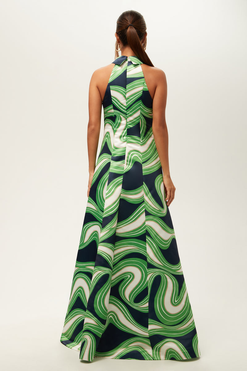 A woman stands facing away in the Official Sleeveless Maxi Dress - Serena Satin, a long halter-neck dress with a bold green and white swirling pattern accented by navy blue. Her hair is pulled back in a low ponytail.