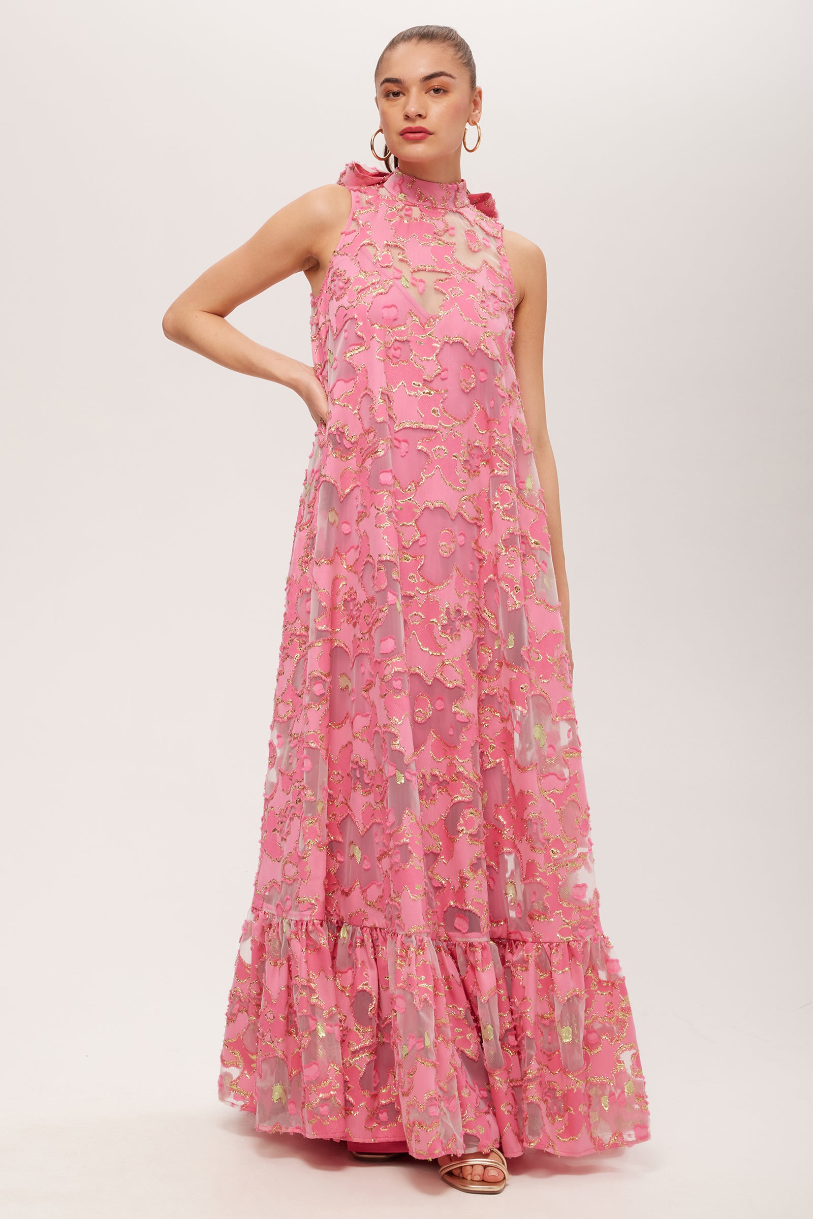 Woman wearing the Leading Dress - Futures Fils Coupe in Love All Pink, a sleeveless floor-length gown with sheer floral overlay, ruffled high neckline and hem. She has hoop earrings and slicked-back hair, against a white background.