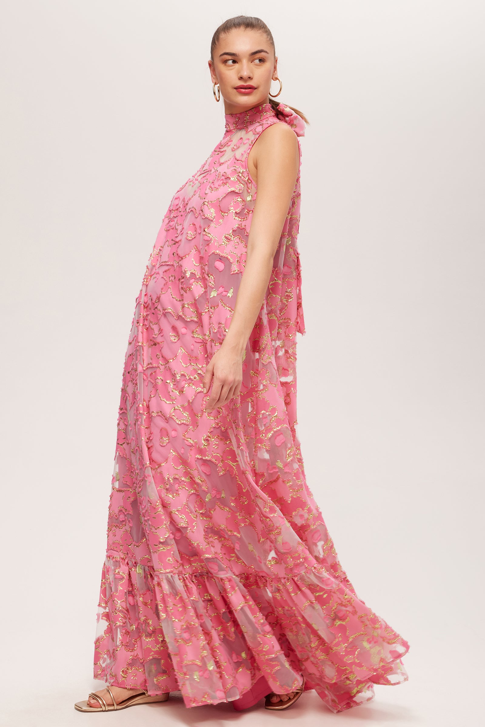 A woman in profile wears the Leading Dress - Futures Fils Coupe: a flowing, sleeveless pink floral dress featuring a high neck and ruffled hem. Her hair is tied back, and she accessorizes with hoop earrings and sandals.