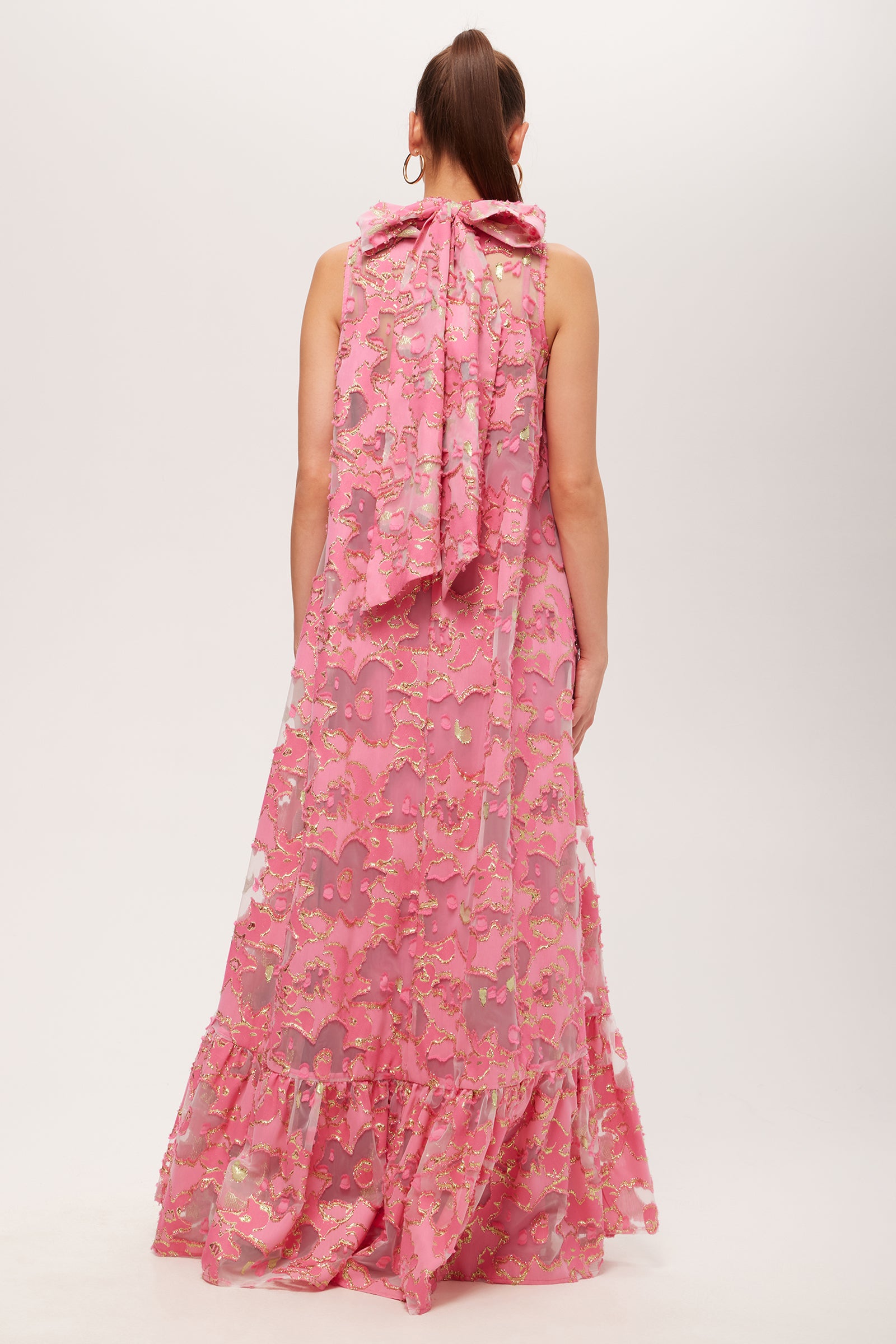 A woman with long brown hair in a ponytail wears the Leading Dress - Futures Fils Coupe, a sleeveless pink floral ankle-length dress with a large back bow and ruffled hem, standing against a plain white background.