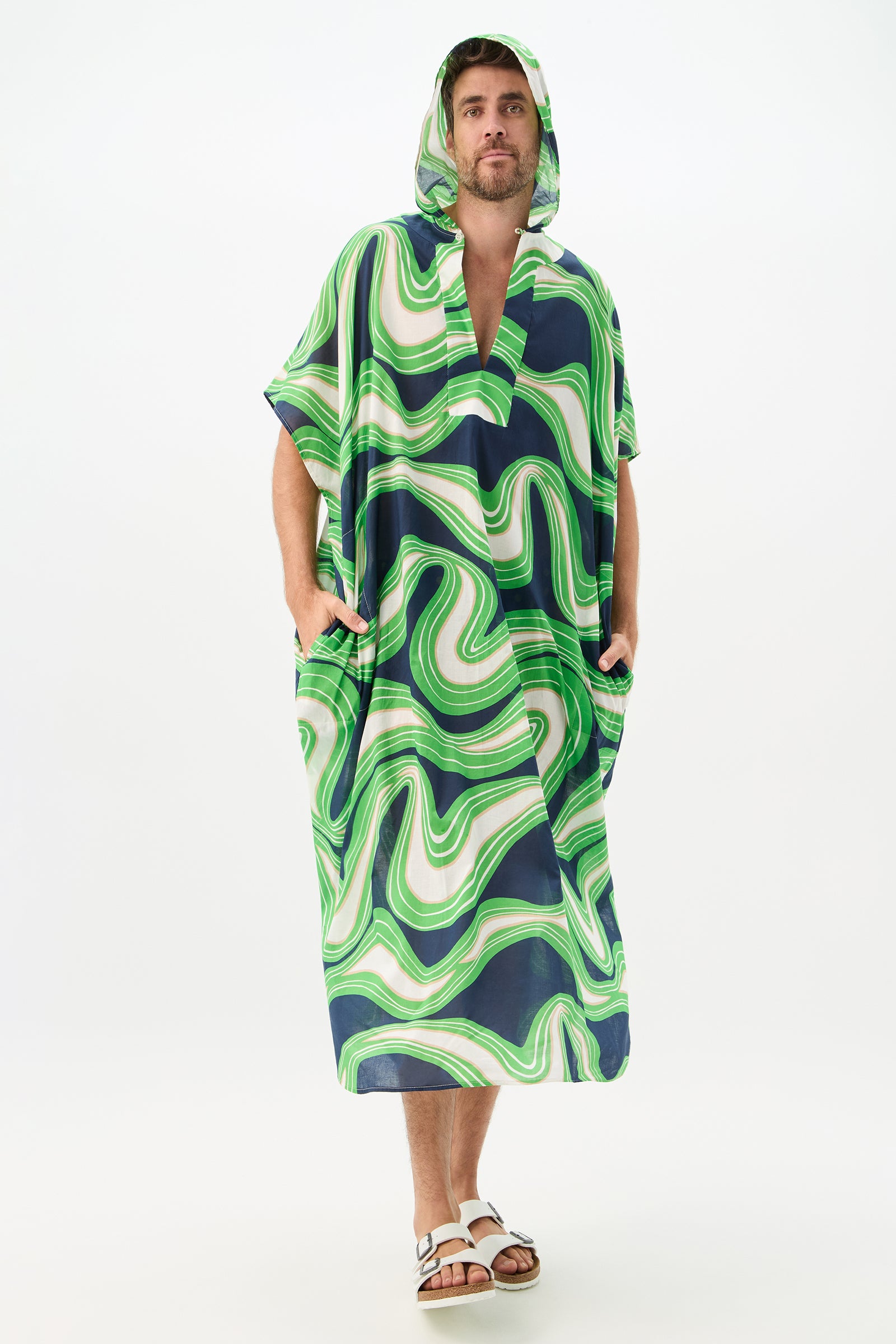 A person stands against a plain white background wearing the Hurley 2 Caftan - Serena Swirl, a loose hooded poncho with swirling green, white, and navy blue patterns, paired with white sandals.