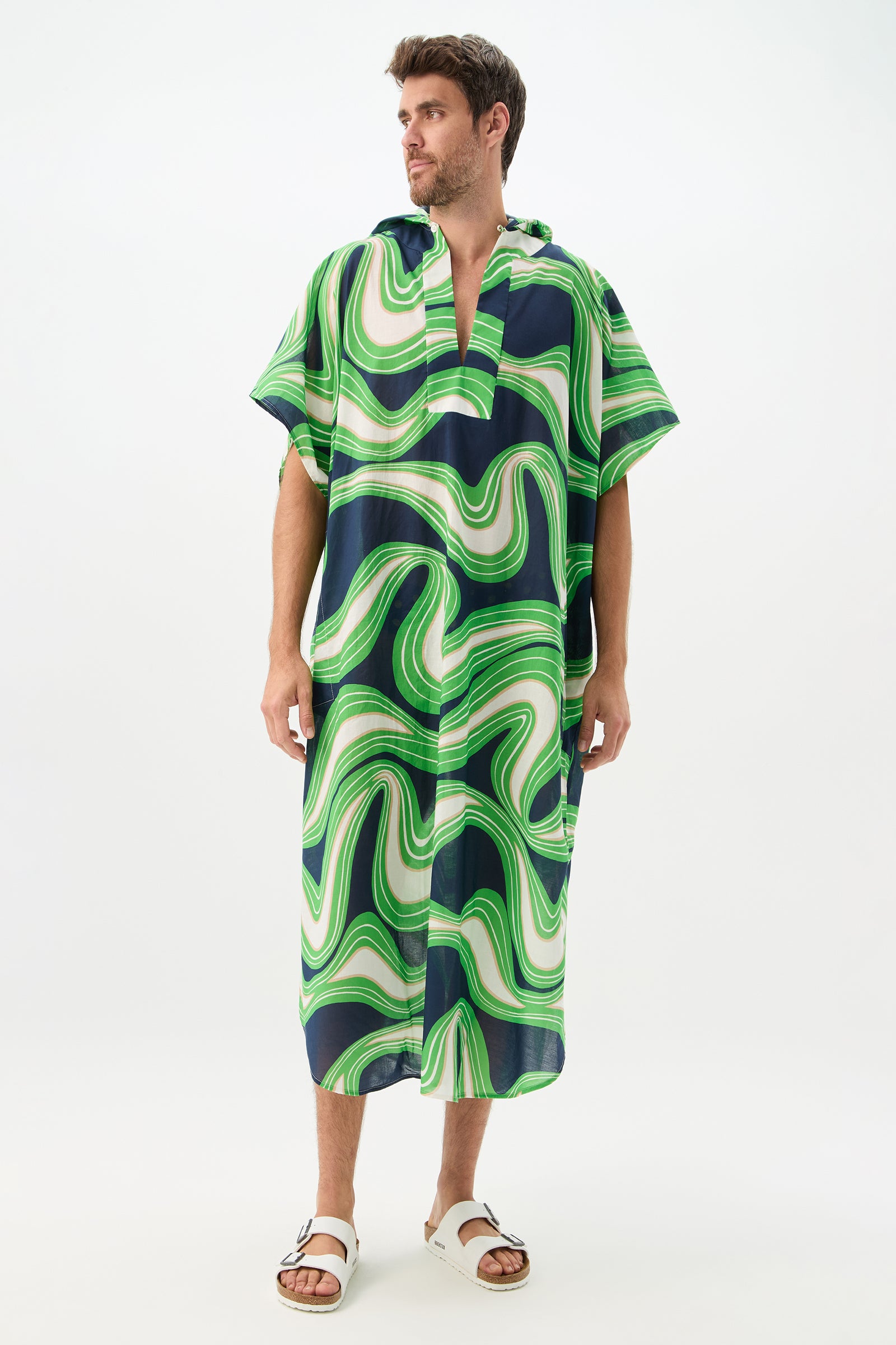 A man stands against a white background wearing the Hurley 2 Caftan - Serena Swirl in Ink, featuring a loose, knee-length fit with bold green, white, and navy swirls. He also wears white sandals and looks slightly to the side.