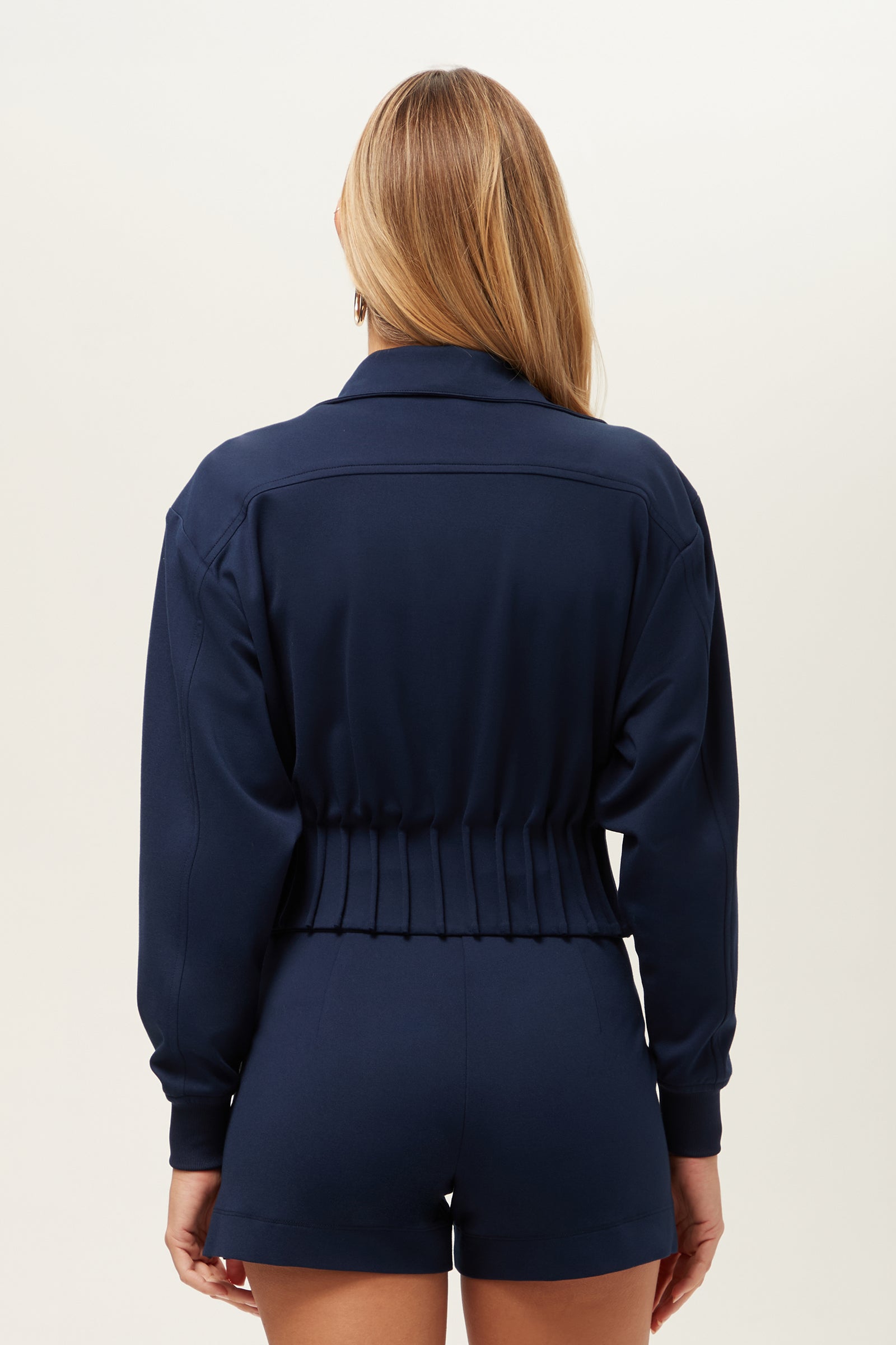A woman with long, straight blonde hair is shown from behind, wearing the navy blue Outrun Jacket - Cozy Ponte and matching high-waisted shorts against a plain light background.