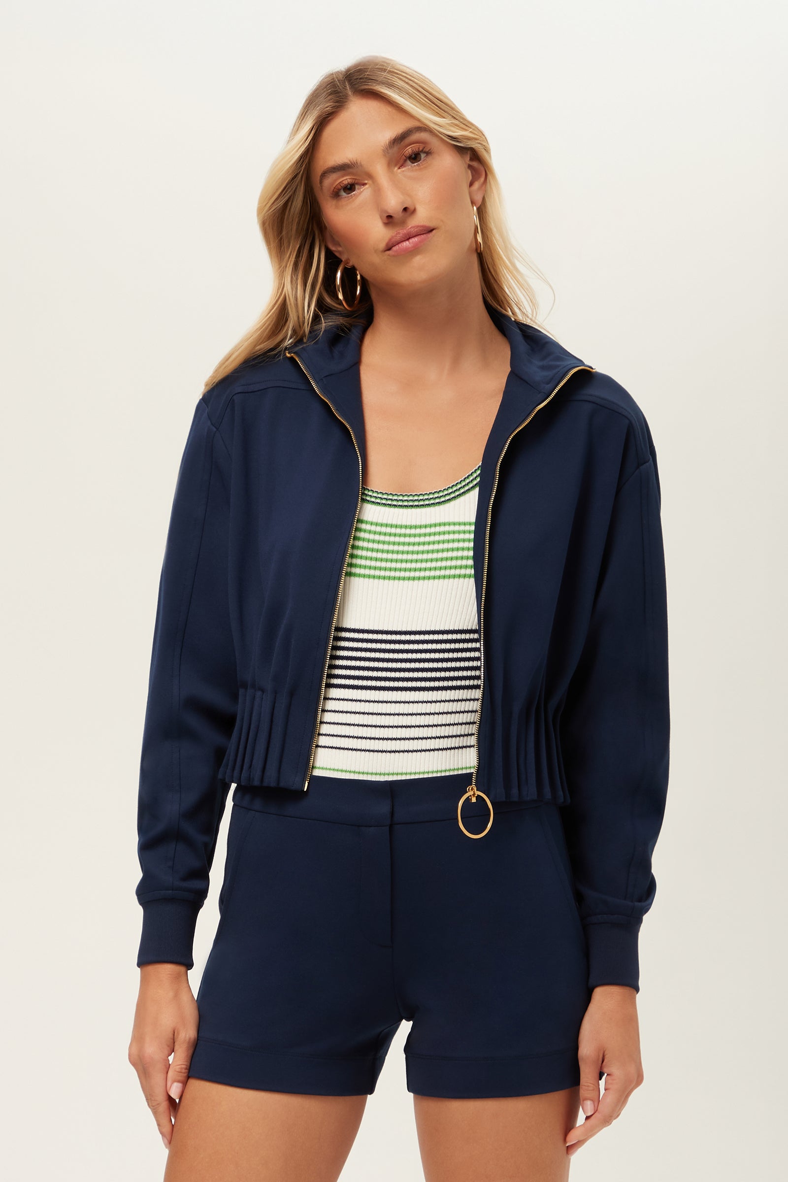 A woman with long blonde hair wears the Outrun Jacket - Cozy Ponte in Ink, paired with matching shorts and a sleeveless green, black, and white striped top. She stands against a light background, looking at the camera.