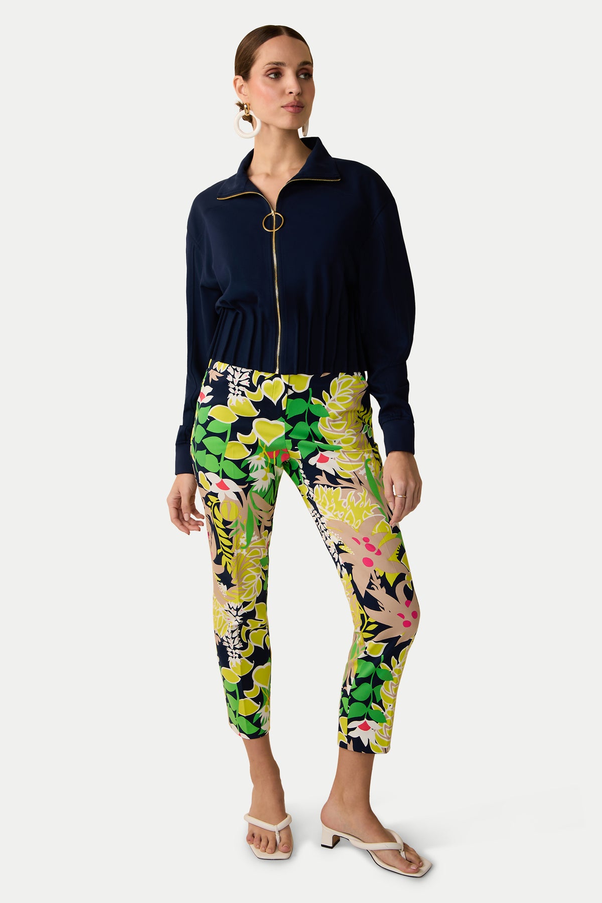 A woman stands wearing a navy zip-up top with a ring zipper and the Aubree 2 Pant - PS Eternal, which features a colorful floral pattern. Her hair is tied back, and she accessorizes with large hoop earrings and white sandals.