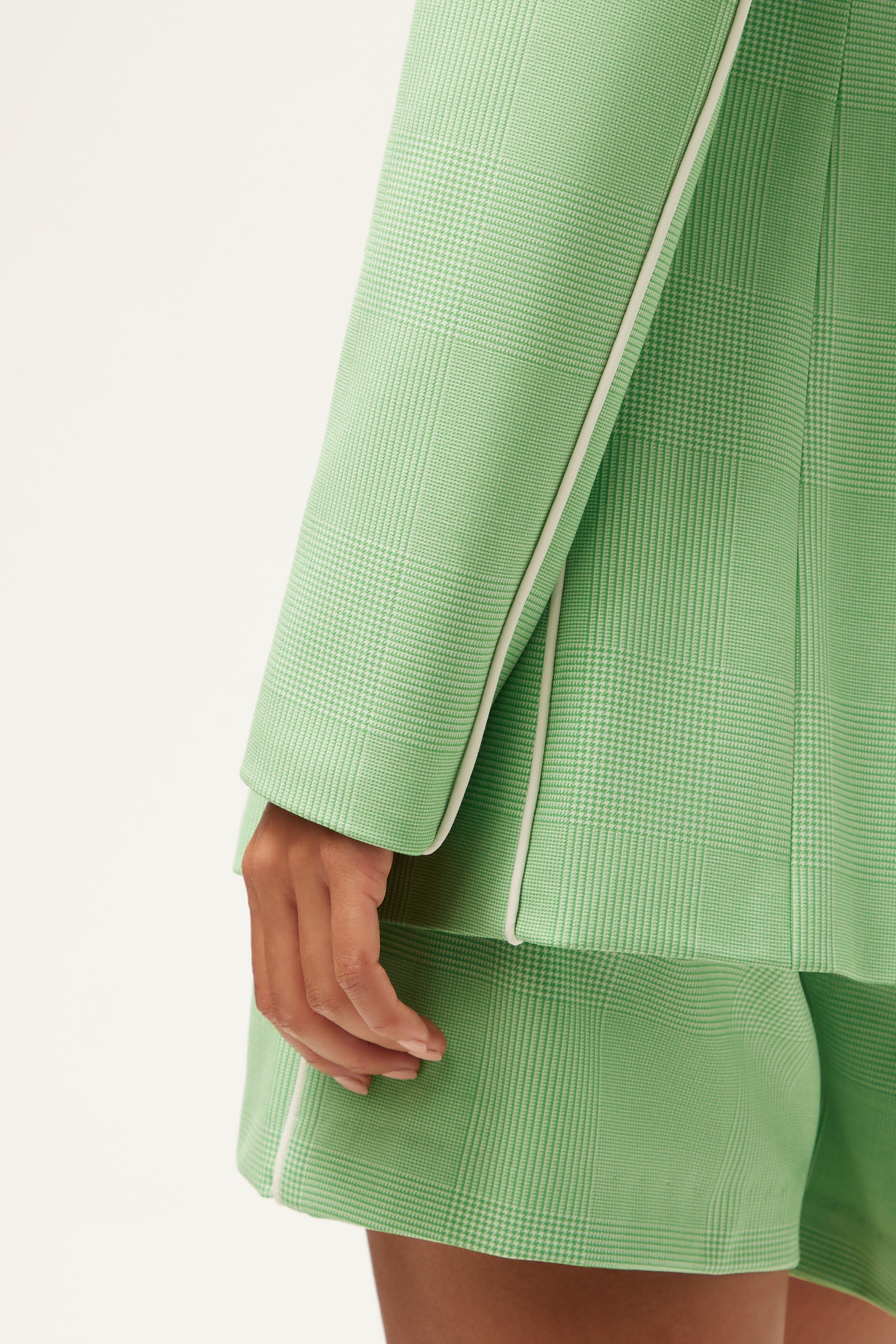 A close-up of a person wearing the Alternates Blazer - Game Time Glen Plaid, a textured light green blazer with white piping, and matching shorts, with their hand resting by their side against a plain background.